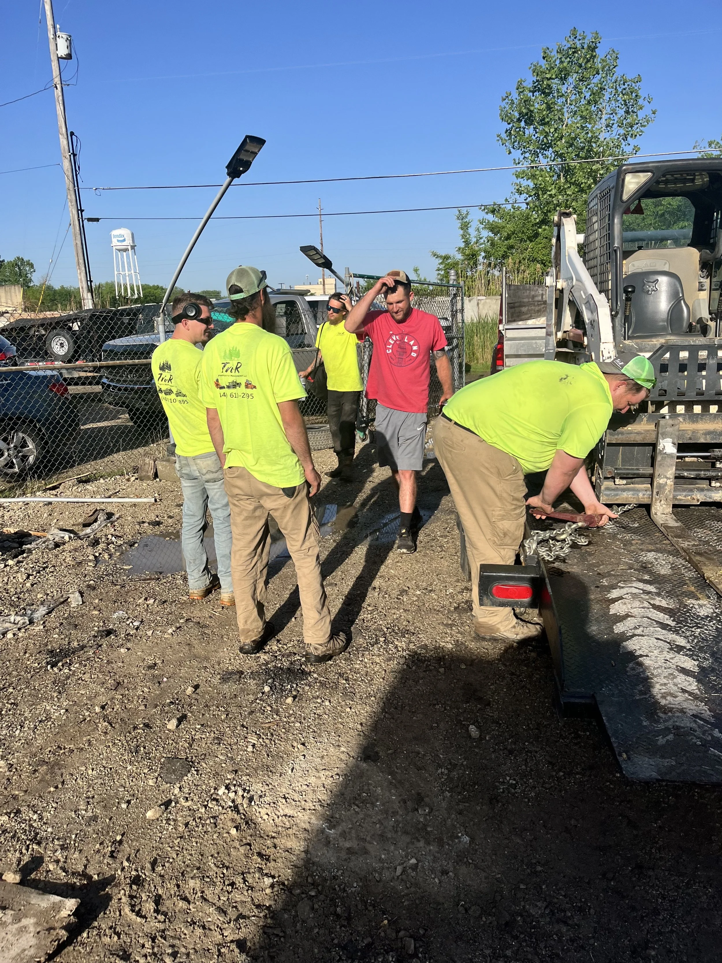Group of construction workers at a worksite with equipment and vehicles, some wearing yellow shirts and safety gear, on a dirt surface under a clear blue sky.