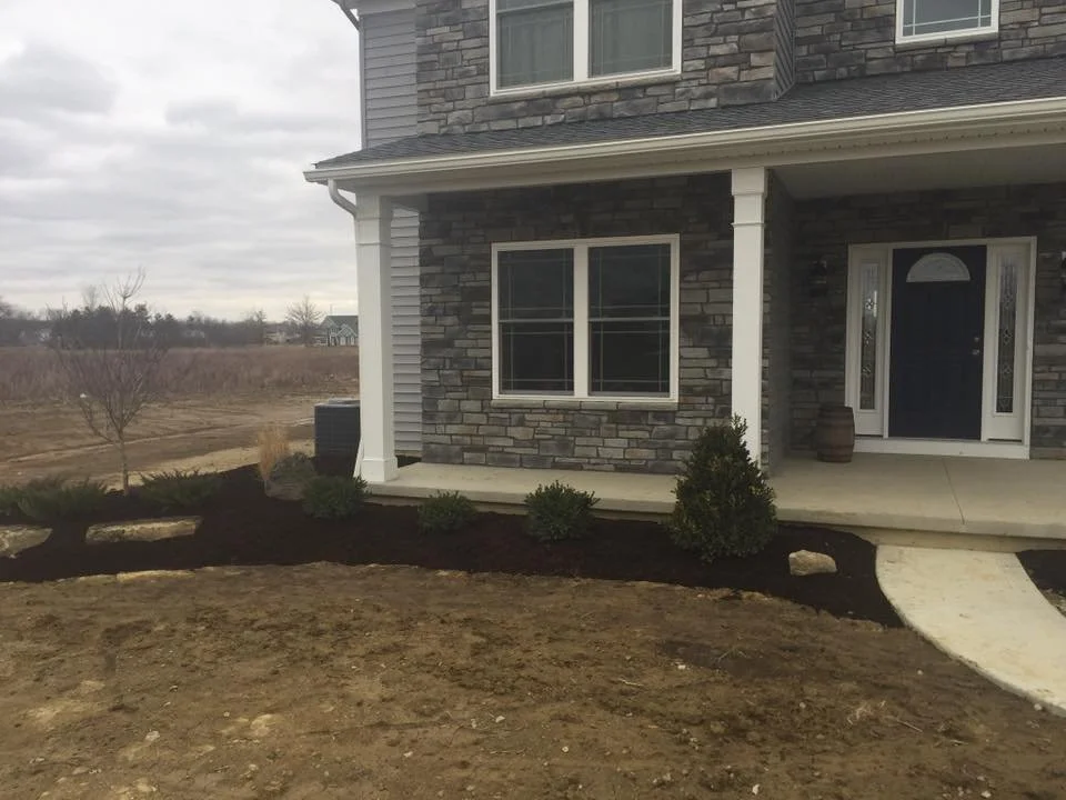 Front porch of a house with new landscaping, including small shrubs and mulch, and a curved walkway leading to the front door.