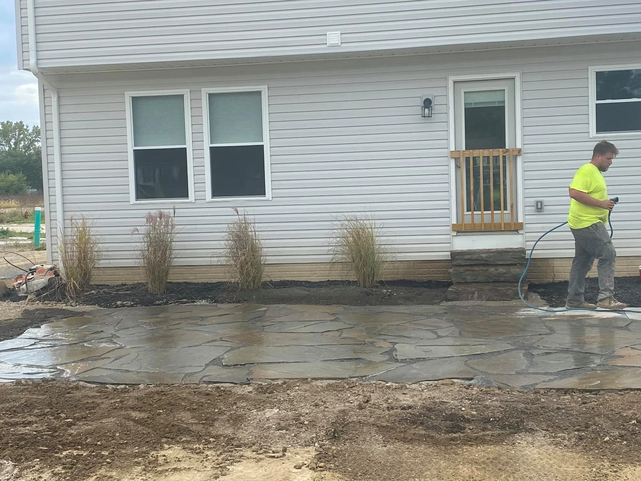 Worker in yellow shirt pouring concrete onto a newly paved stone patio in front of a white house.