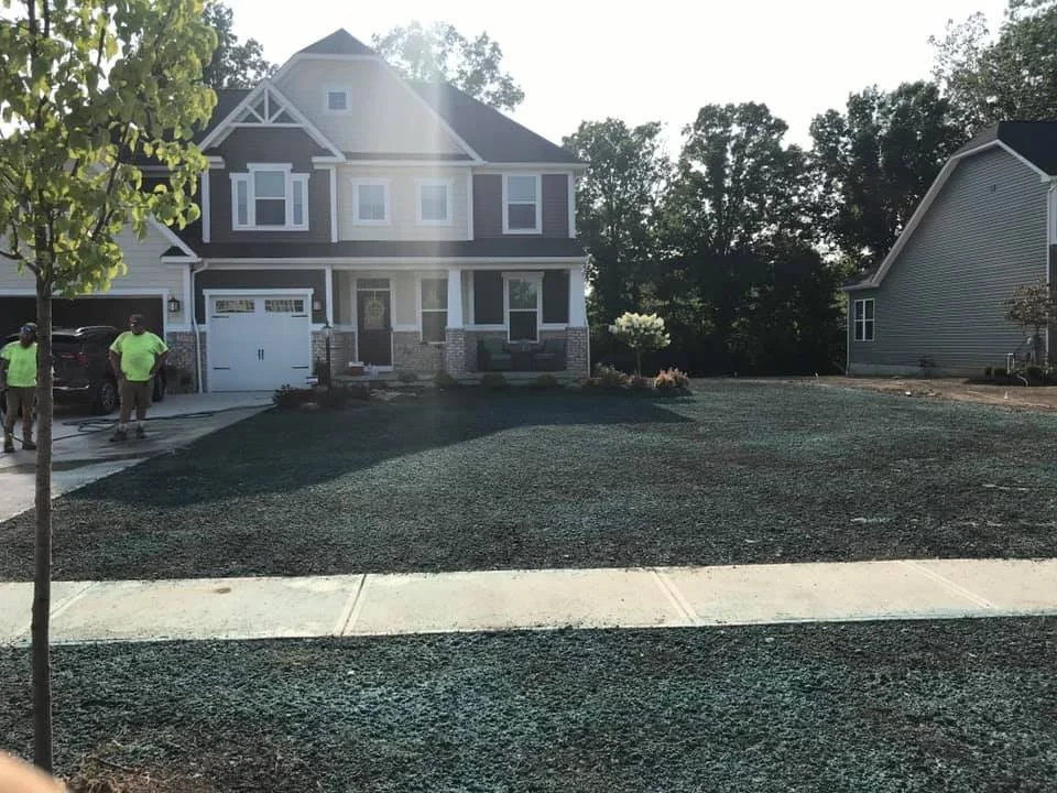 Newly landscaped front yard with fresh black mulch, sidewalk, and a two-story house with a garage and trees in the background.