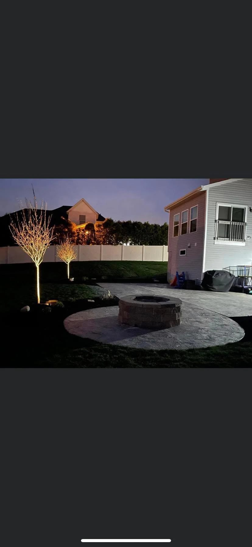Nighttime backyard scene with two lit trees, a stone fire pit, a white house with a balcony, and neighboring houses in the background under a clear sky.
