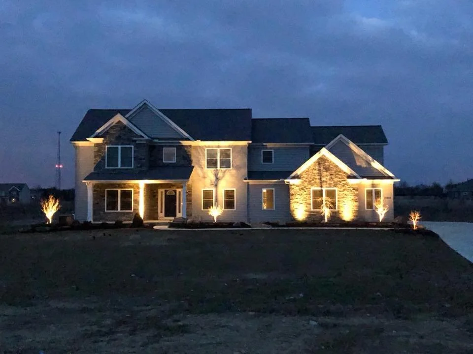 A large two-story house illuminated at night with exterior lighting, manicured lawn, and small landscaped trees.
