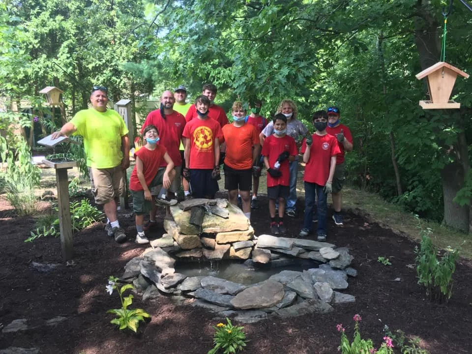 Group of children and adults standing around a small artificial pond with a rock waterfall in a garden, some wearing masks and gloves, with birdhouses hanging from trees and a garden sign visible.