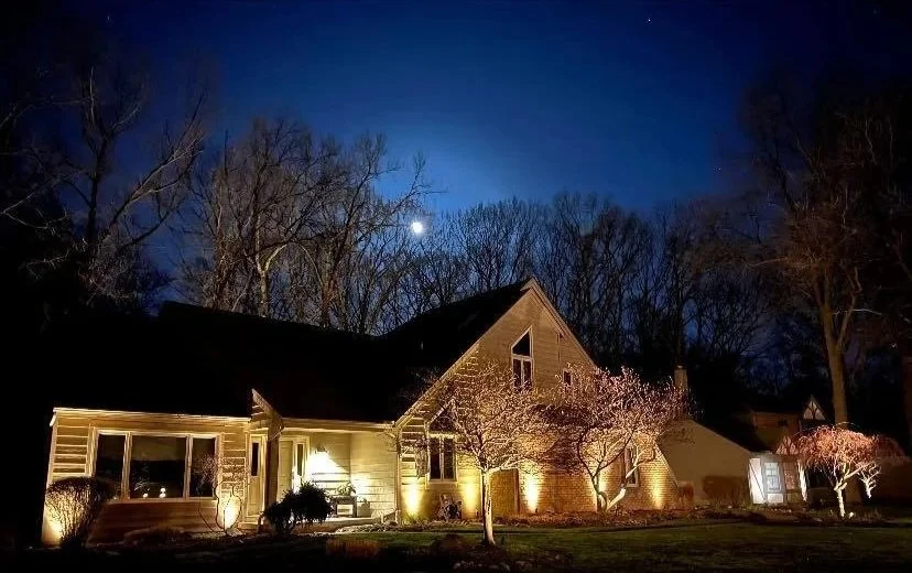 Nighttime view of a house illuminated with exterior lights, surrounded by trees with pink blossoms, under a dark sky with a bright moon.