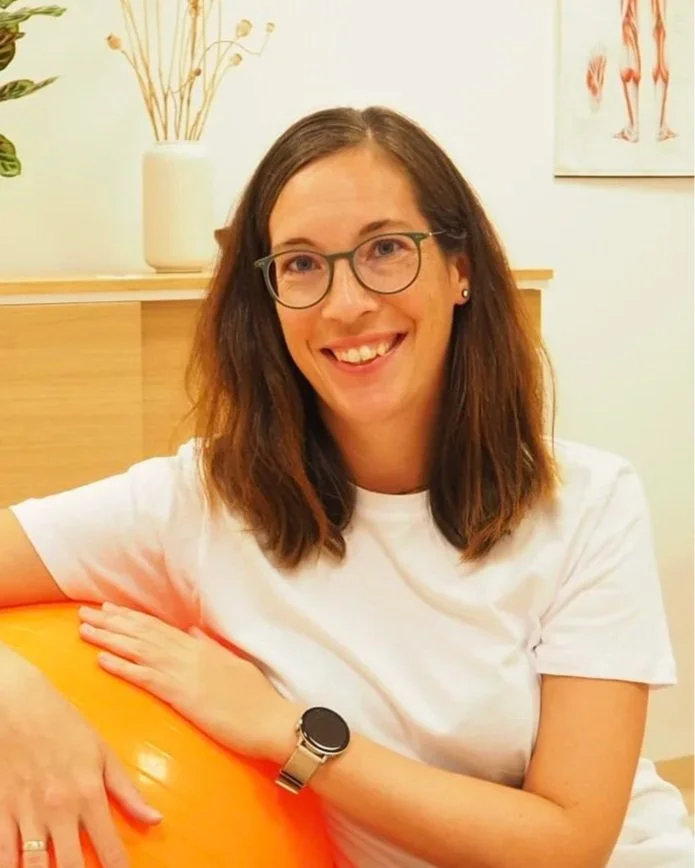 Eine lächelnde Frau mit Brille, braunen Haaren, weißem T-Shirt und einem schwarzen Armband hält einen orangefarbenen Ball in einem Raum mit Pflanzen und anatomischen Postern im Hintergrund. Die Frau ist Sabine Wieser, BSc. Physiotherapeutin.