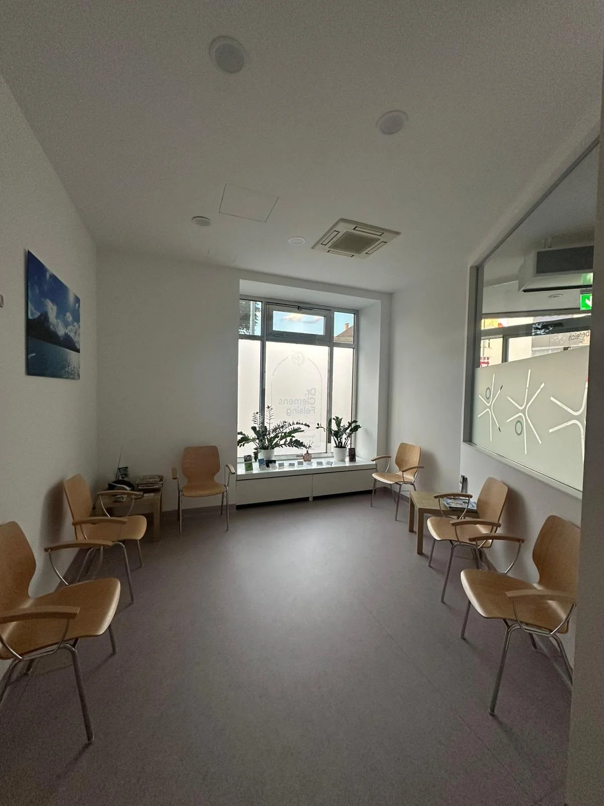 Wartezimmer mit Stühlen, Fenster mit Pflanzen, Wandbild, helle Wände, neutrales Design, Tageslicht.