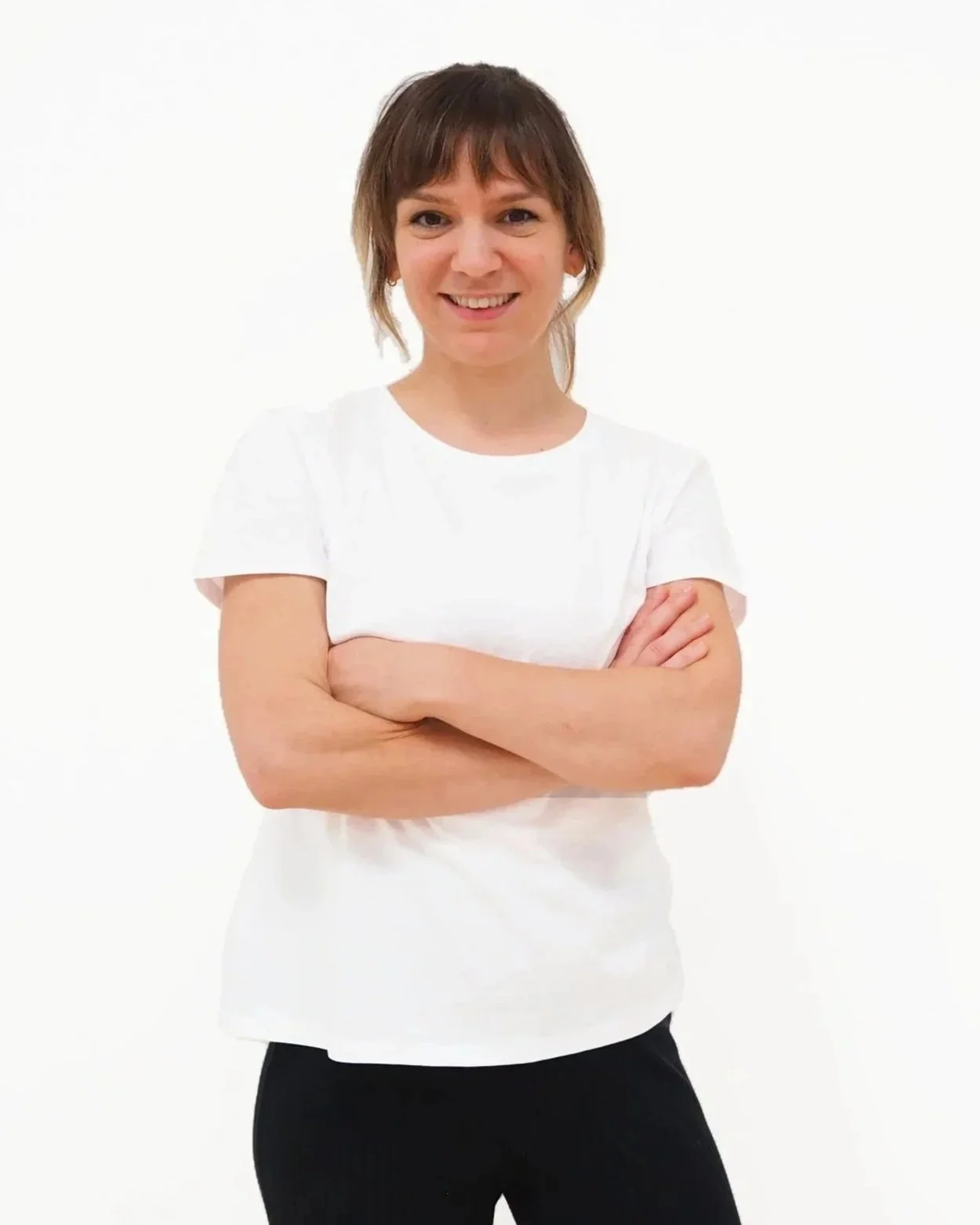 Junge Frau mit kurzen braunen Haaren, trägt ein weißes T-Shirt und schwarze Hosen, steht vor weißem Hintergrund, lächelt und hat die Arme verschränkt. Die Frau ist Katharina Widhalm, BSc. Physiotherapeutin und Kinderphysiotherapeutin.