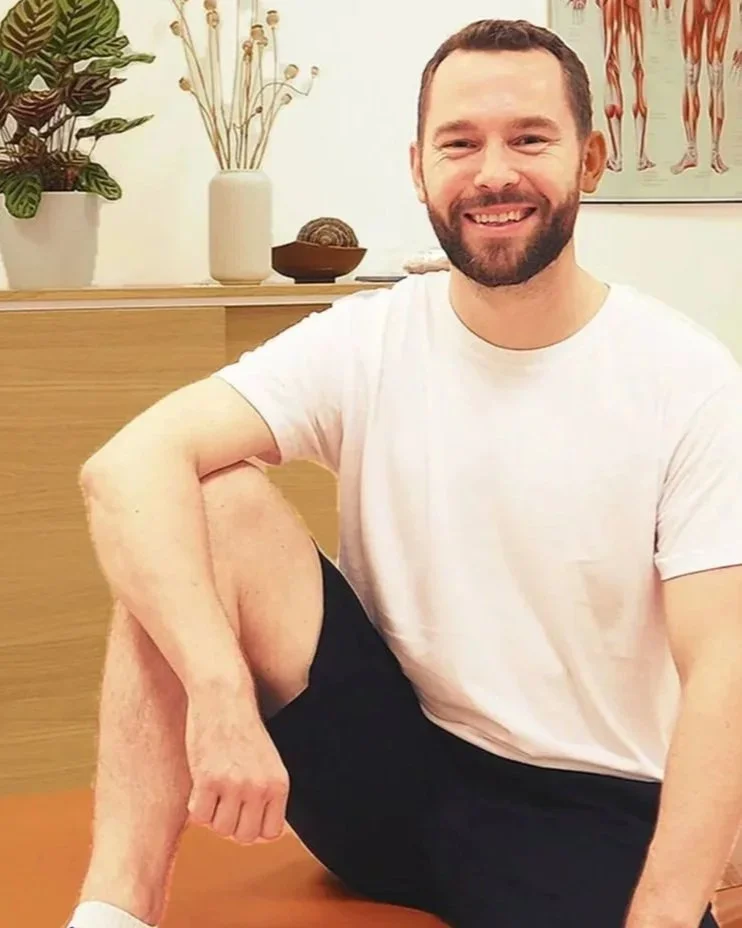 Ein lachender Mann mit Bart in einem weißen T-Shirt sitzt auf dem Boden, mit einem Bein über das andere gebeugt, in einem Raum mit Pflanzen und anatomischen Postern. Der Mann ist Manuel Walzel, BSc. Physiotherapeut und Manualtherapeut.