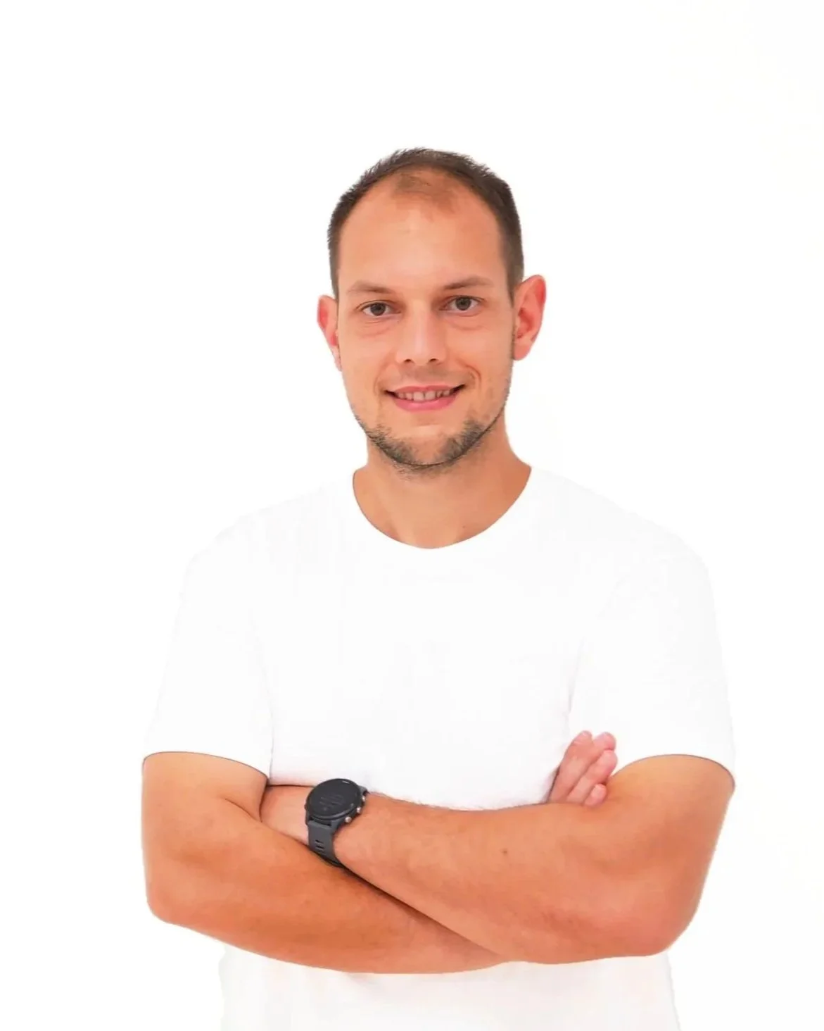 Junger Mann mit kurzen Haaren, legerem weißen T-Shirt, trägt eine schwarze Armbanduhr, steht vor weißem Hintergrund, lächelt mit verschränkten Armen. Der Mann ist Nicolas Oberleitner, BSc. Physiotherapeut und Sportphysiotherapeut.