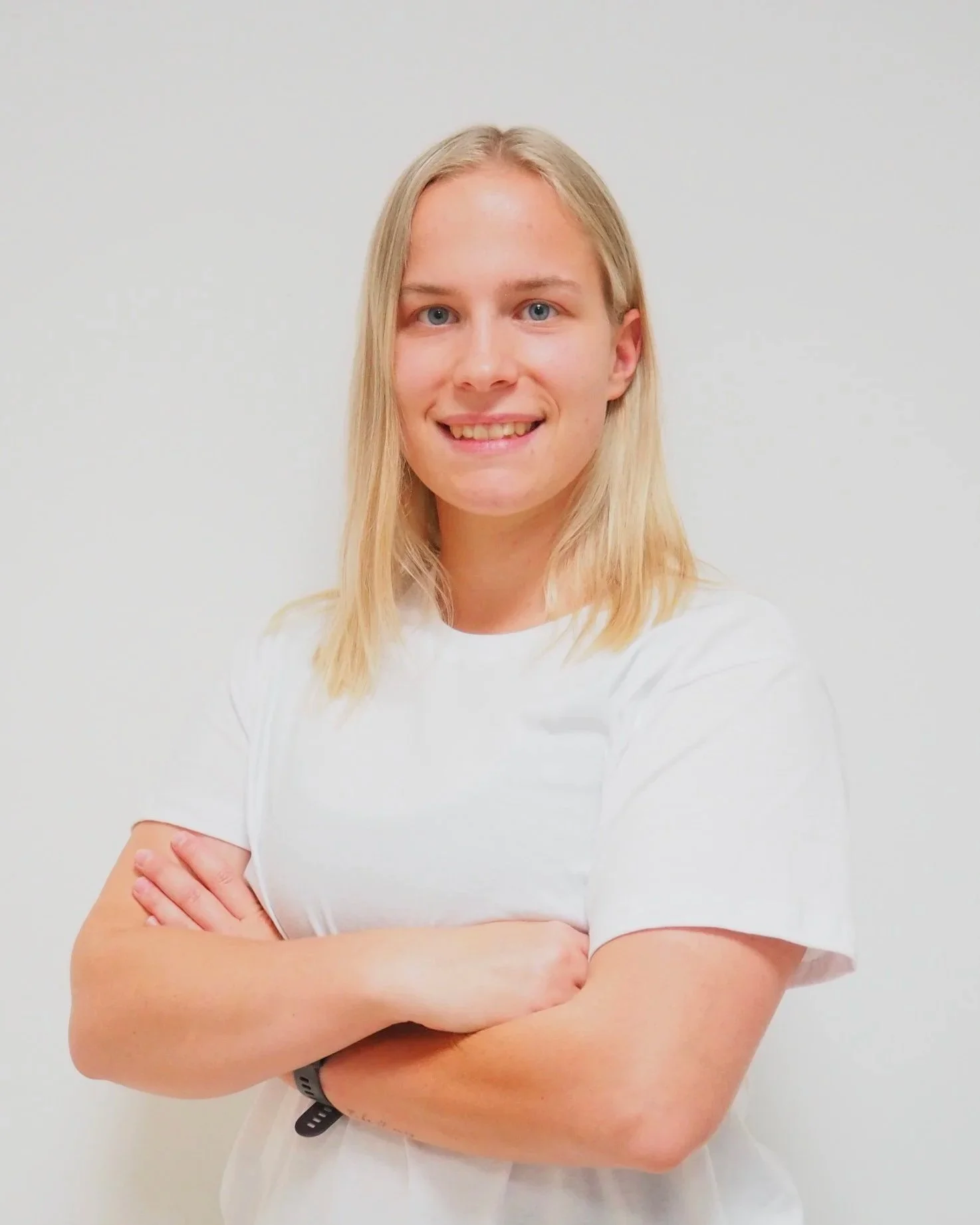 Junge Frau mit blonden Haaren und blauen Augen, trägt weißes T-Shirt, lächelt, steht vor weißem Hintergrund, die Arme sind verschränkt. Die Frau ist Elena Oberleitner, BSc. Physiotherapeutin und Sportphysiotherapeutin.