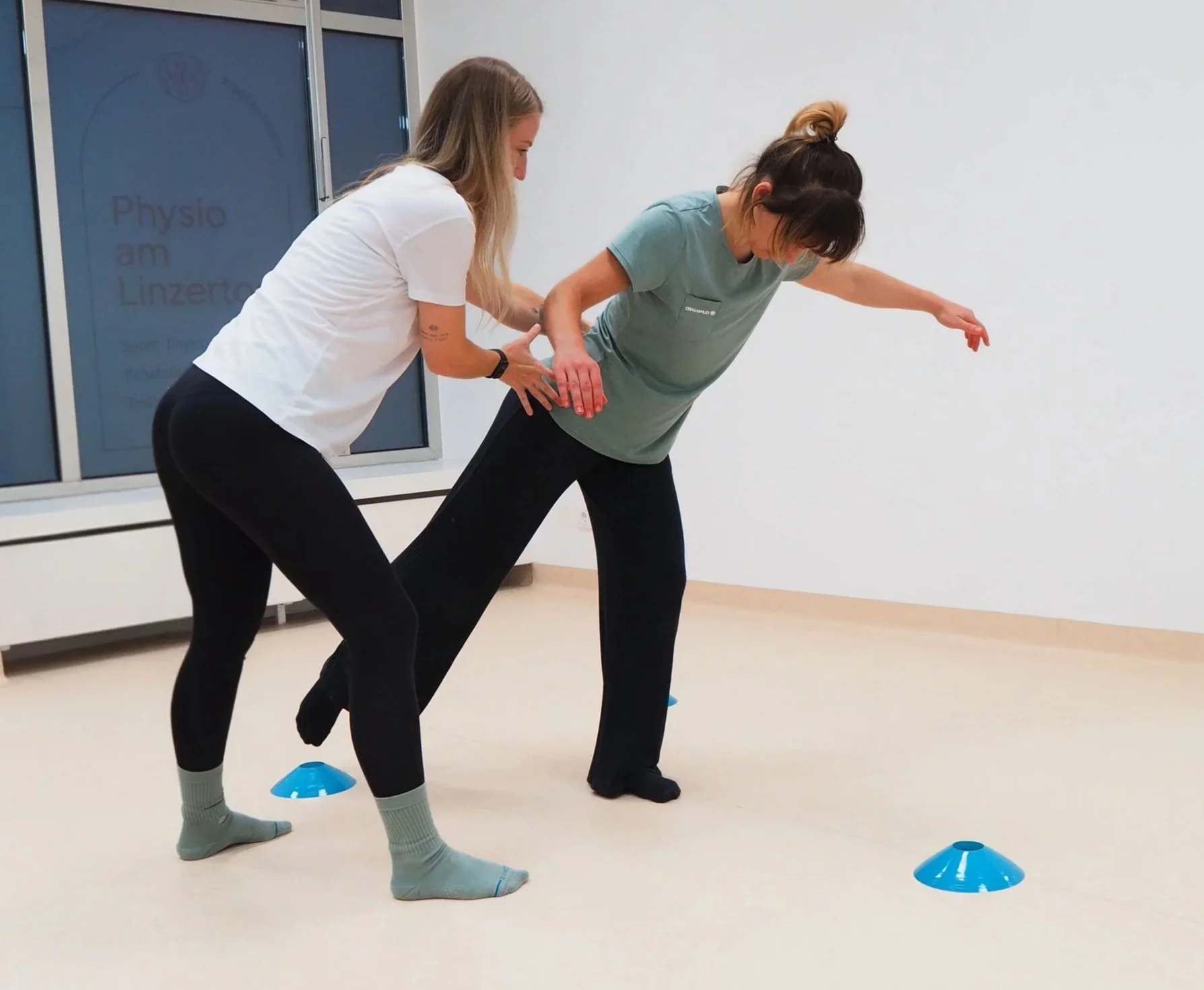 Zwei Frauen üben zusammen Balance auf einem Bein, während sie an einer Physiotherapie-Übung teilnehmen, innerhalb eines Raumes mit weißen Wänden und blauen Markierungshütchen auf dem Boden.