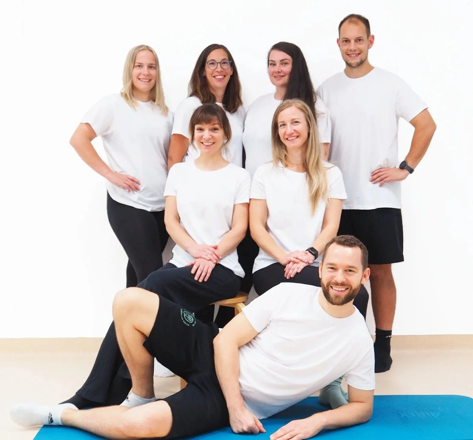 Gruppe von sieben Menschen, fünf Frauen und zwei Männer, in weißen T-Shirts, posieren vor weißem Hintergrund. Einige sitzen, andere stehen, alle lächeln. Das Team von Physio am Linzertor.