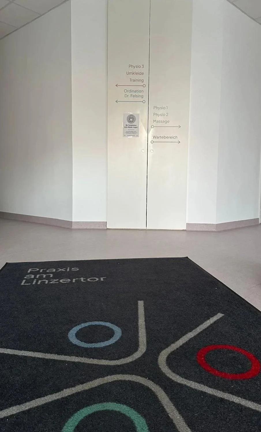 Der Flur in einer medizinischen Praxis mit schwarzen Teppich, auf dem 'Praxis am Linzertor' steht, und weißen Wänden mit Wegweiserschildern zu Physio- und Trainingsräumen, sowie Wartebereich.