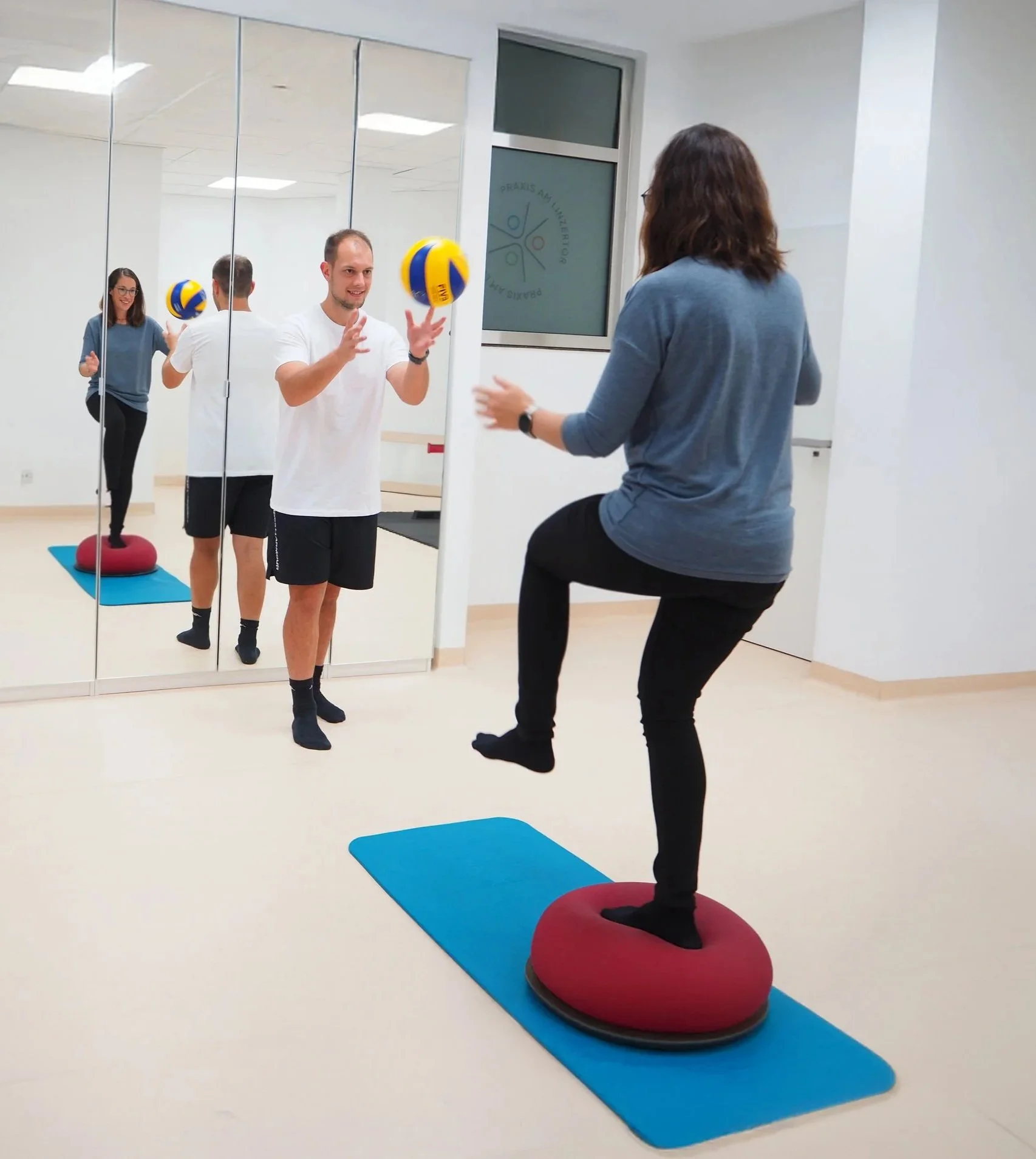 Zwei Menschen üben Balancetraining auf einer rot umwickelten Trampolin-Variante im Trainingsraum. Eine Person steht auf dem Trampolin, die andere wirft ihr einen Ball zu. Nicolas Oberleitner Physiotherapeut und Sportphysiotherapeut
