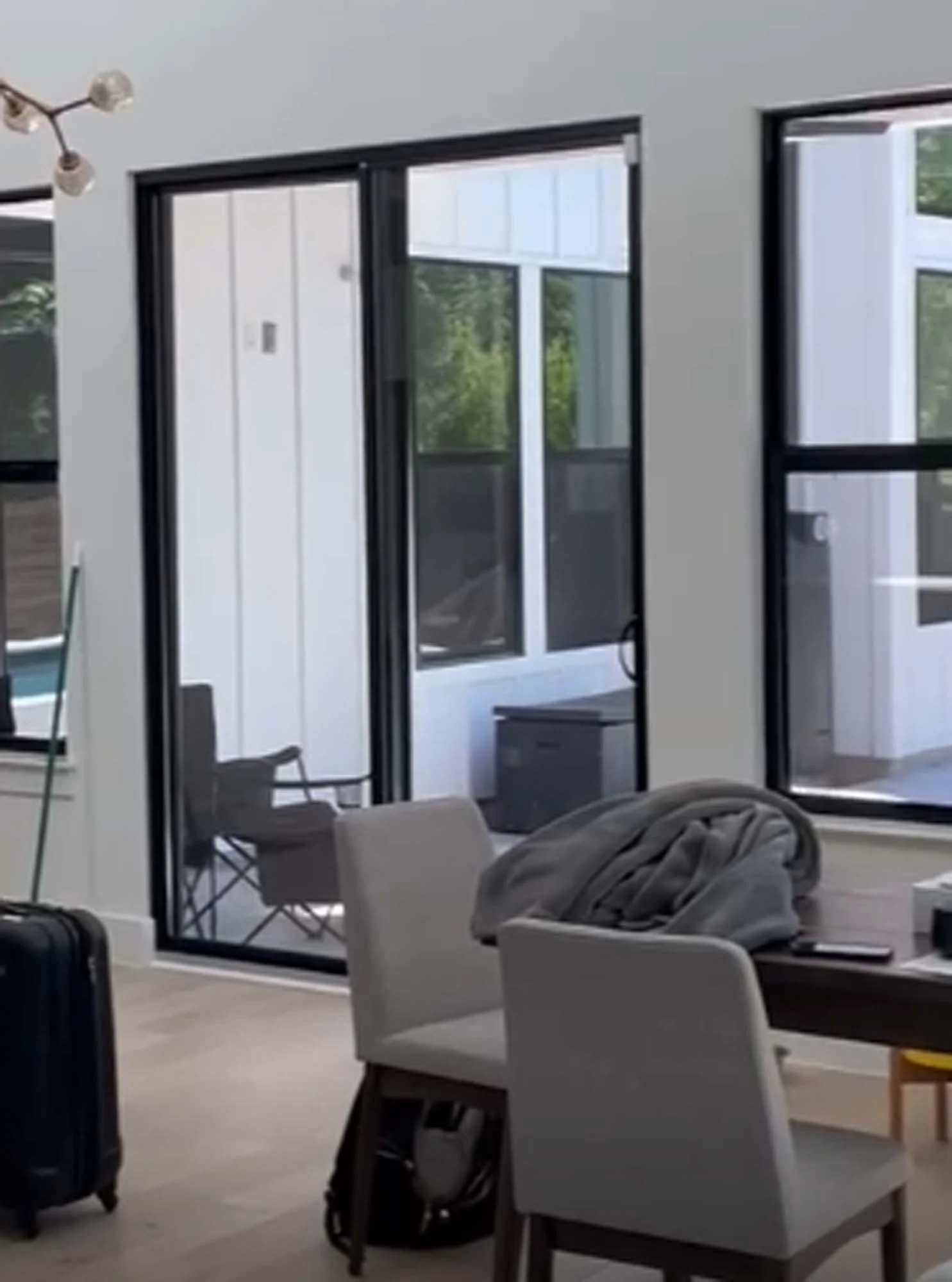 Interior of a modern house with large glass sliding doors, a dining table with a blanket and phone on top, two chairs, a suitcase, and visible outdoor balcony with chairs and trees.