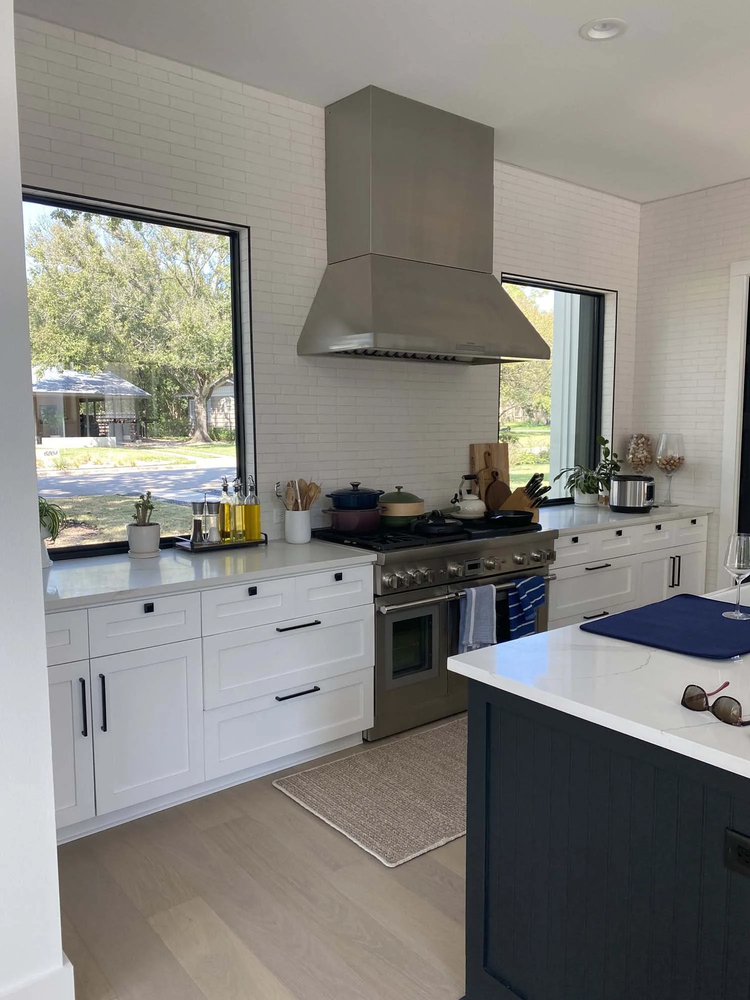 Modern kitchen with white cabinets, stainless steel stove and range hood, black cabinet on island, and windows showing a sunny outdoor scene with trees.