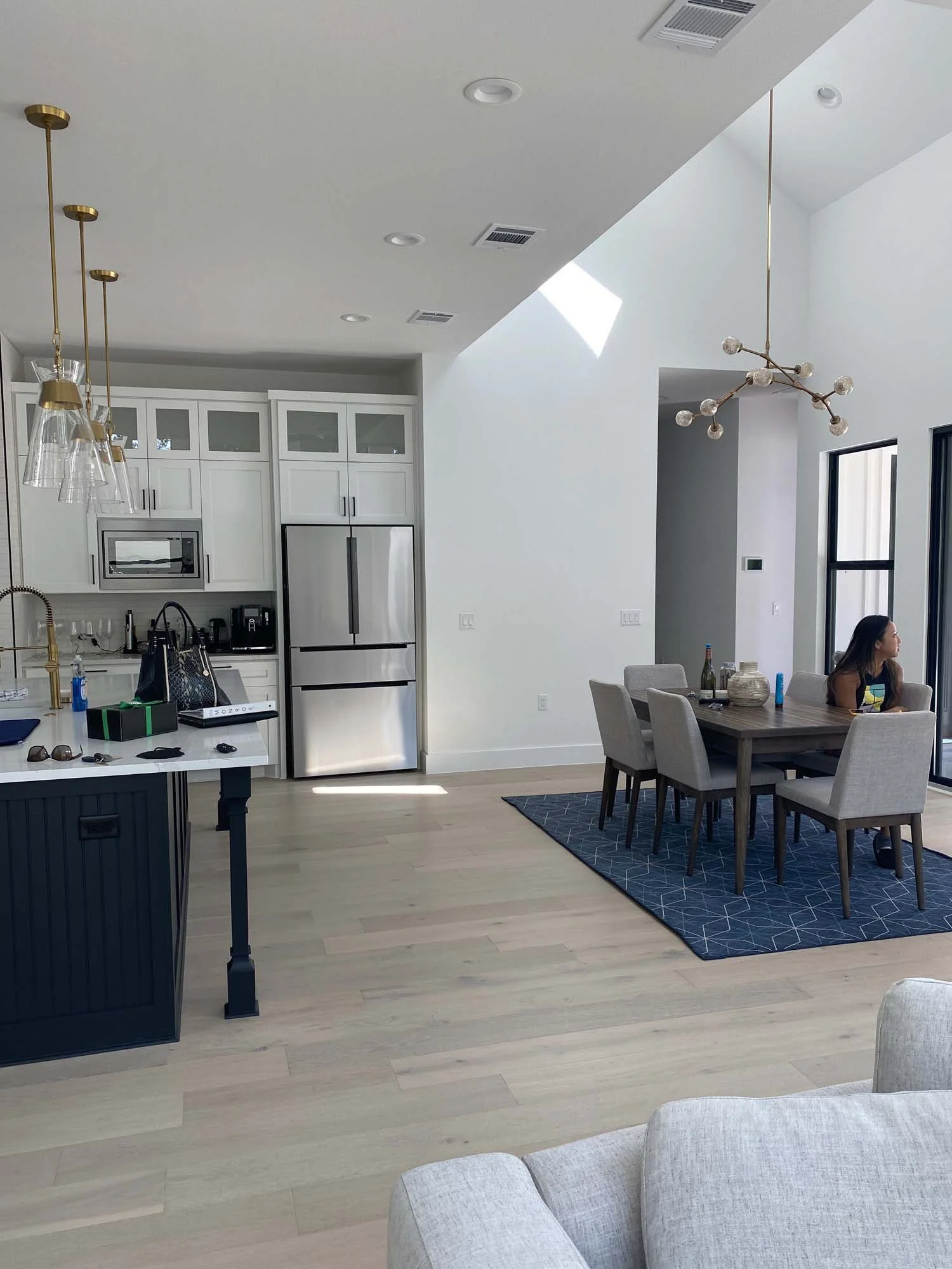 An open plan kitchen and dining area with white cabinets, stainless steel refrigerator, light hardwood floors, and a modern chandelier. A woman is sitting at a dining table with beige chairs, and a blue geometric-patterned rug. There is a sliding gla