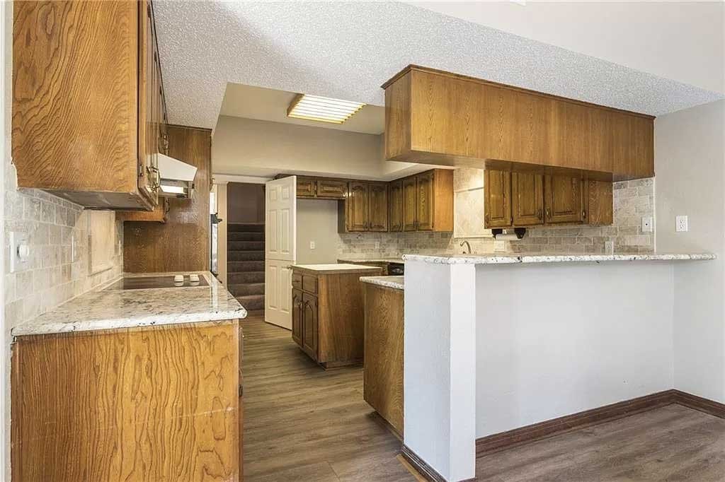 Kitchen with wooden cabinets, granite countertops, brick backsplash, white walls, and wooden floor. There is a doorway leading to stairs in the background.