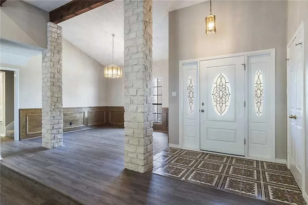 Entryway of a house with a white front door with decorative glass panels, tiled flooring in front of the door, and wood flooring in the adjacent room. There are stone columns, wainscoting, and hanging light fixtures.