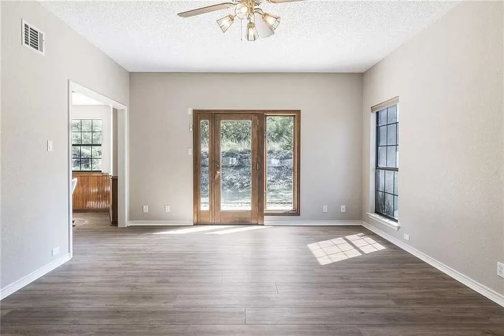 Empty living room with wood floor, white walls, a ceiling fan, and sliding glass door leading outside, with sunlight streaming in.