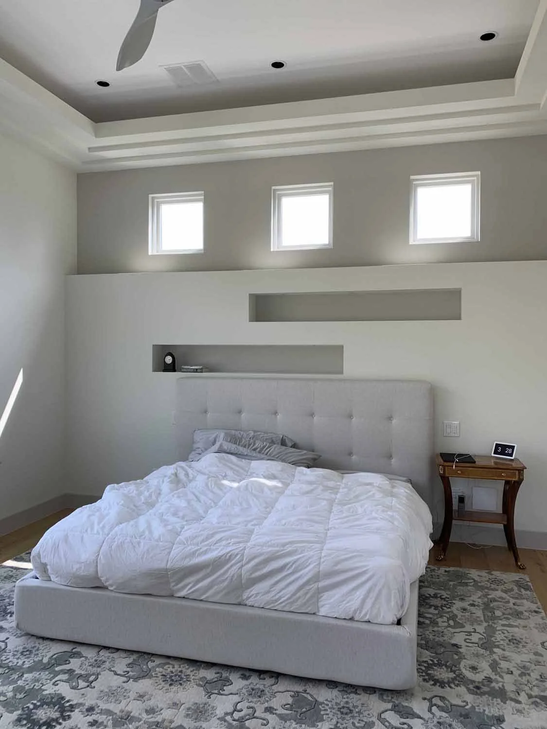 Modern bedroom with a white upholstered bed, a wooden nightstand with a digital clock, and three small windows near the ceiling, with beige walls and a gray and white patterned rug.