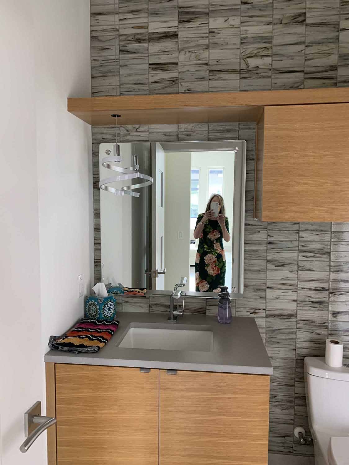Bathroom mirror reflecting a woman in a floral dress, standing in a brightly lit room, with a vanity with a faucet, soap dispenser, folded towels, tissue box, and a toilet with a roll of toilet paper.