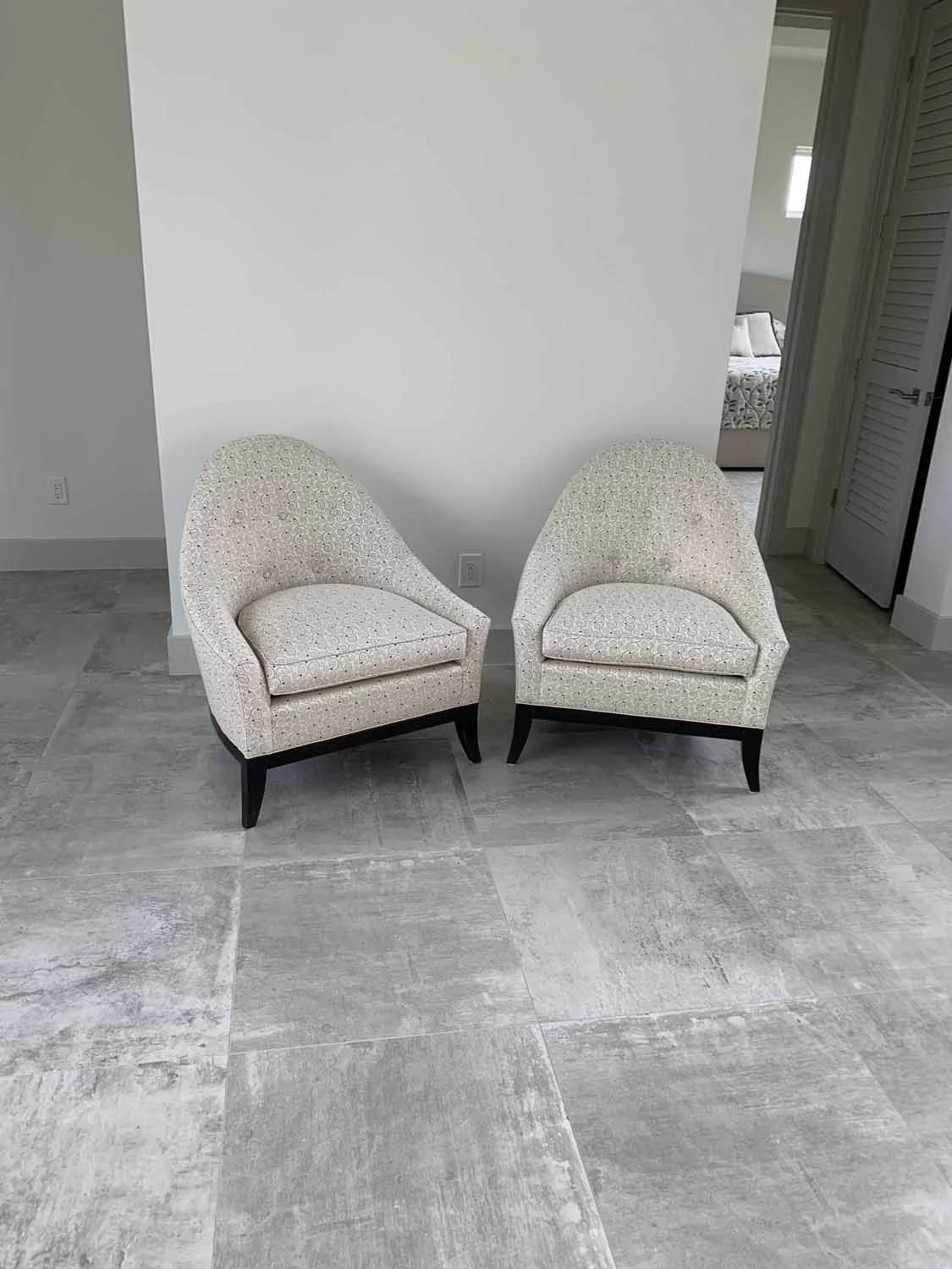 Two identical beige upholstered armchairs with dark wooden legs are placed side by side against a white wall in a room with rectangular gray tile flooring.