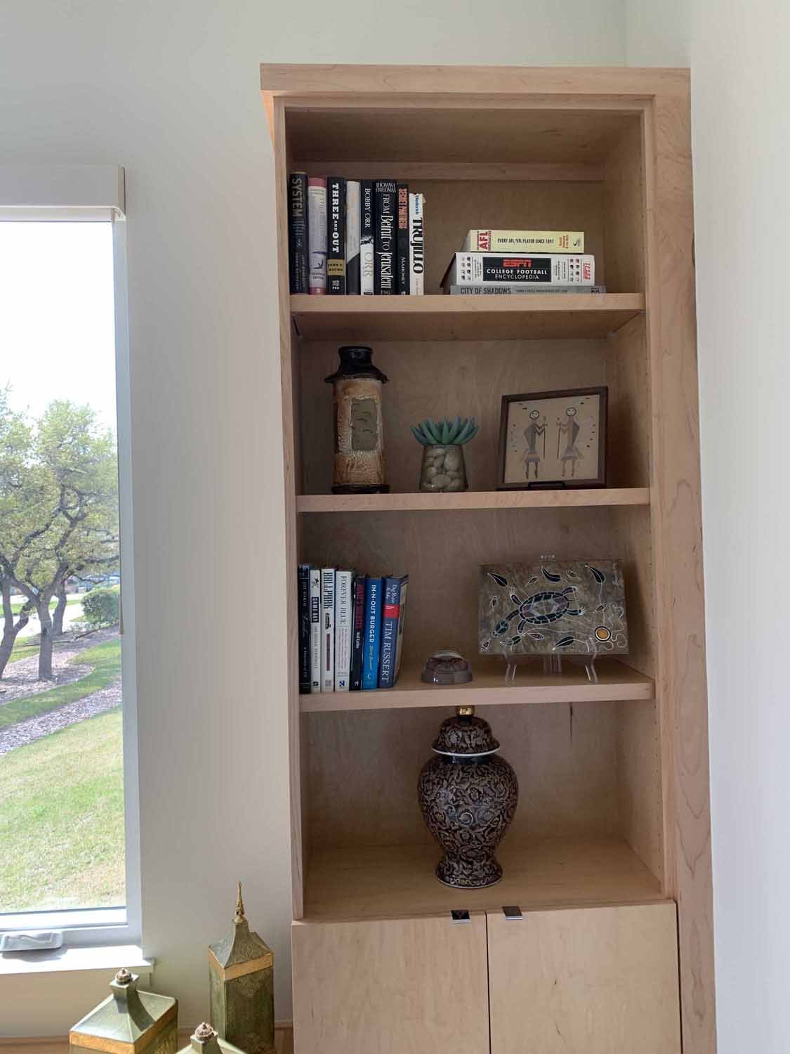 Wooden bookshelf with five shelves holding books, decorative objects, and artwork, positioned next to a window with trees outside.