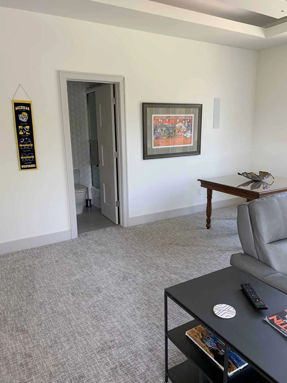 Interior view of a living room with a doorway leading to a bathroom. The room has a carpeted floor, a wall-mounted frame, a side table with decorative items, a grey armchair, and a small black table with books, a remote, and decorative pieces.