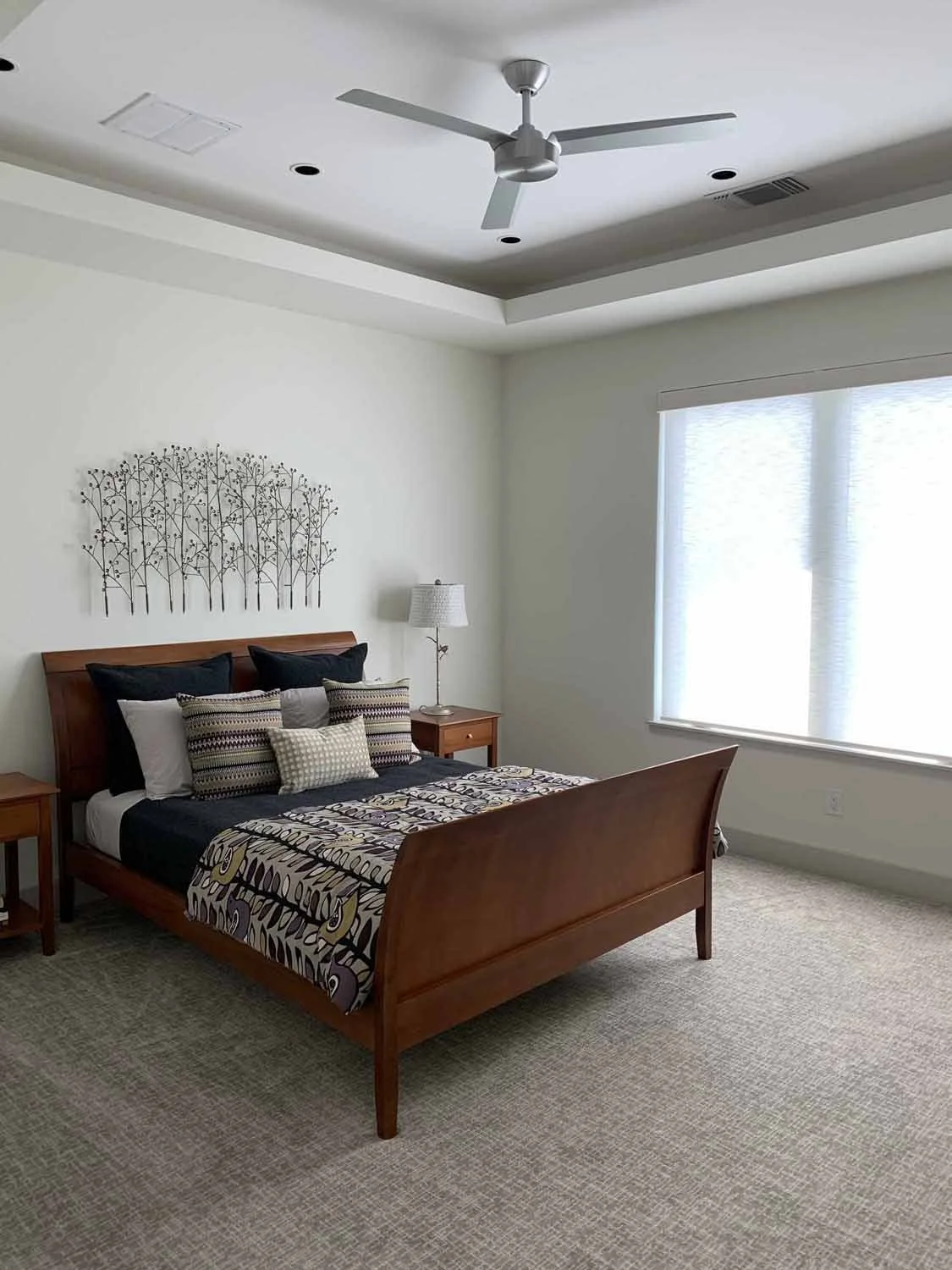 A bedroom with a wooden bed frame, black and patterned pillows, a decorative metal wall art above the headboard, a wooden nightstand with a lamp, a large window with white blinds, a ceiling fan, and light-colored carpet.