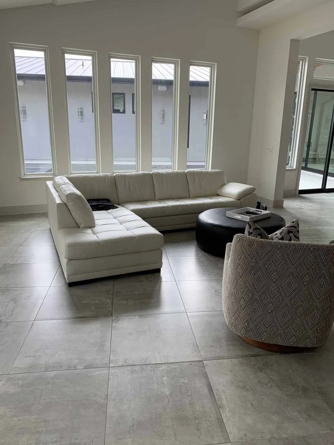 Living room with a large white sectional sofa, a round black coffee table, patterned armchair, and large windows with a view of the neighboring building.
