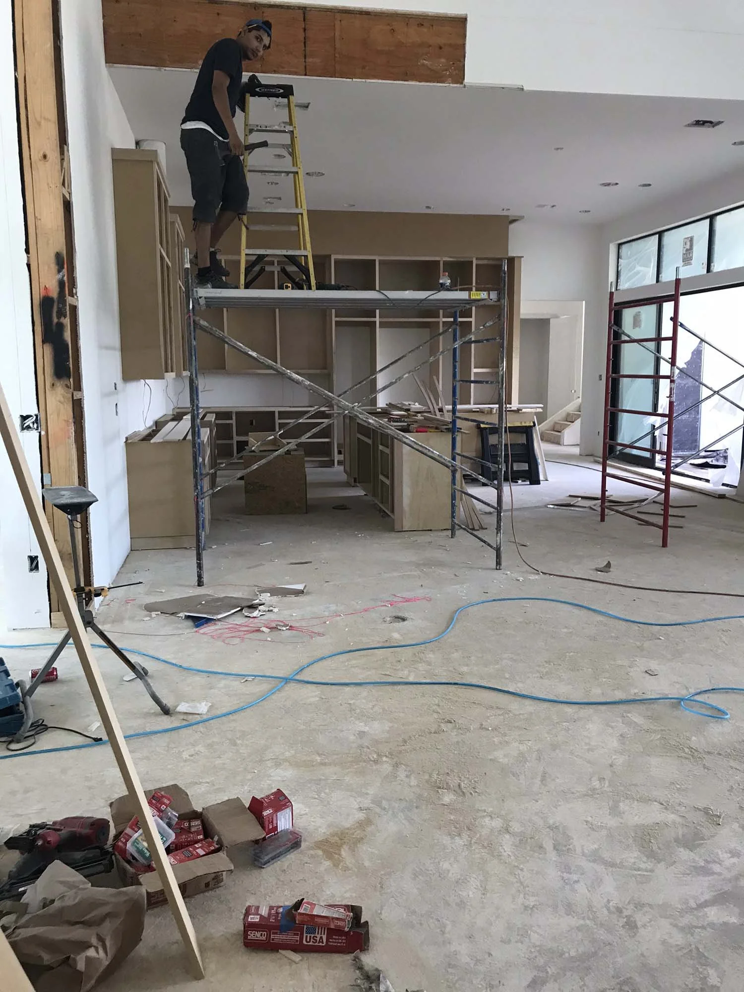 Interior of a building under construction with a person standing on scaffolding, working on the ceiling. Construction tools and materials are scattered on the floor.