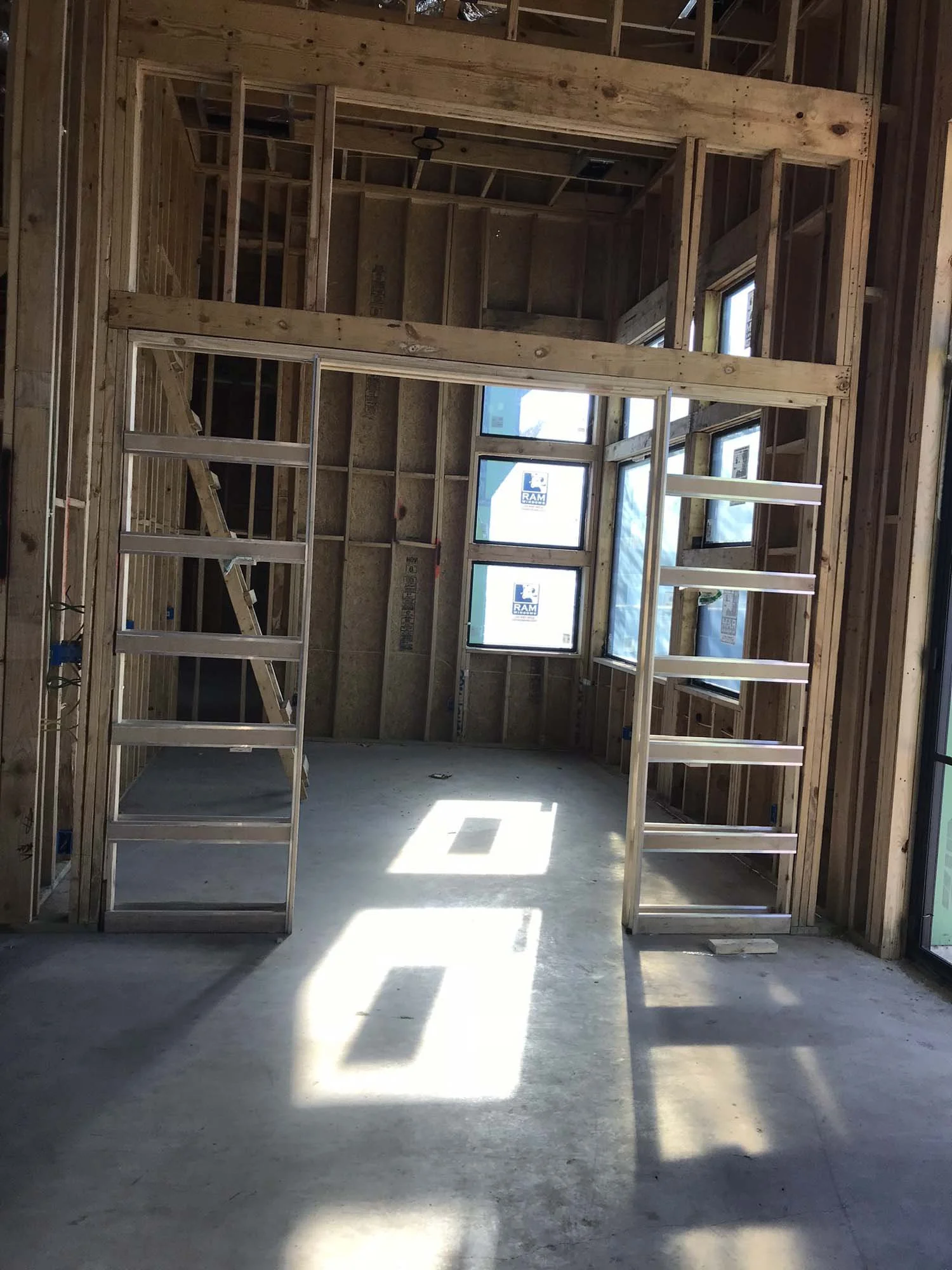 Interior of a house under construction, showing exposed wooden framing, a loft area, and sunlight casting geometric shadows on the concrete floor.