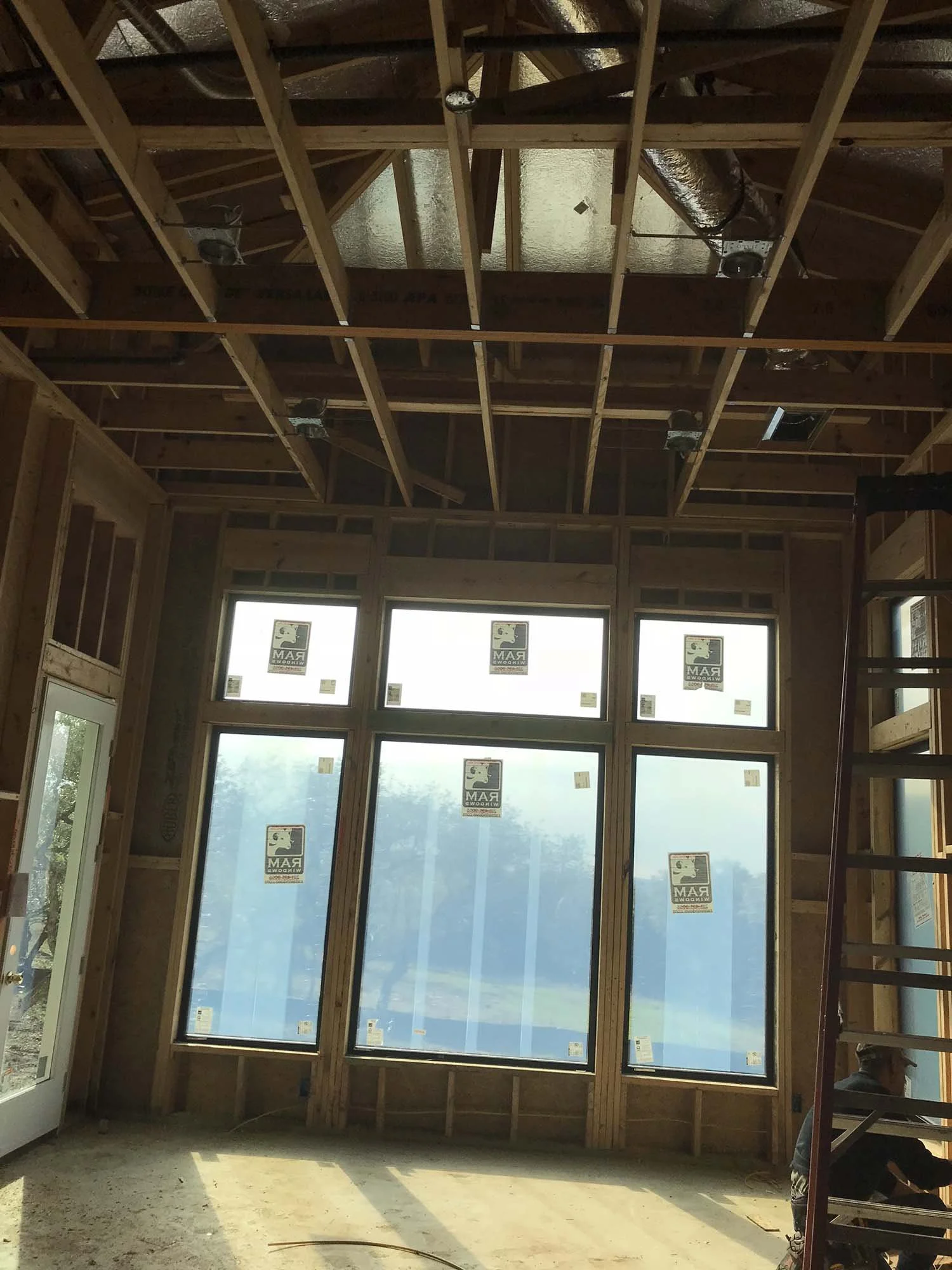 Interior of a house under construction showing framed walls, large window with stickers, and exposed ceiling with wooden beams and ductwork.