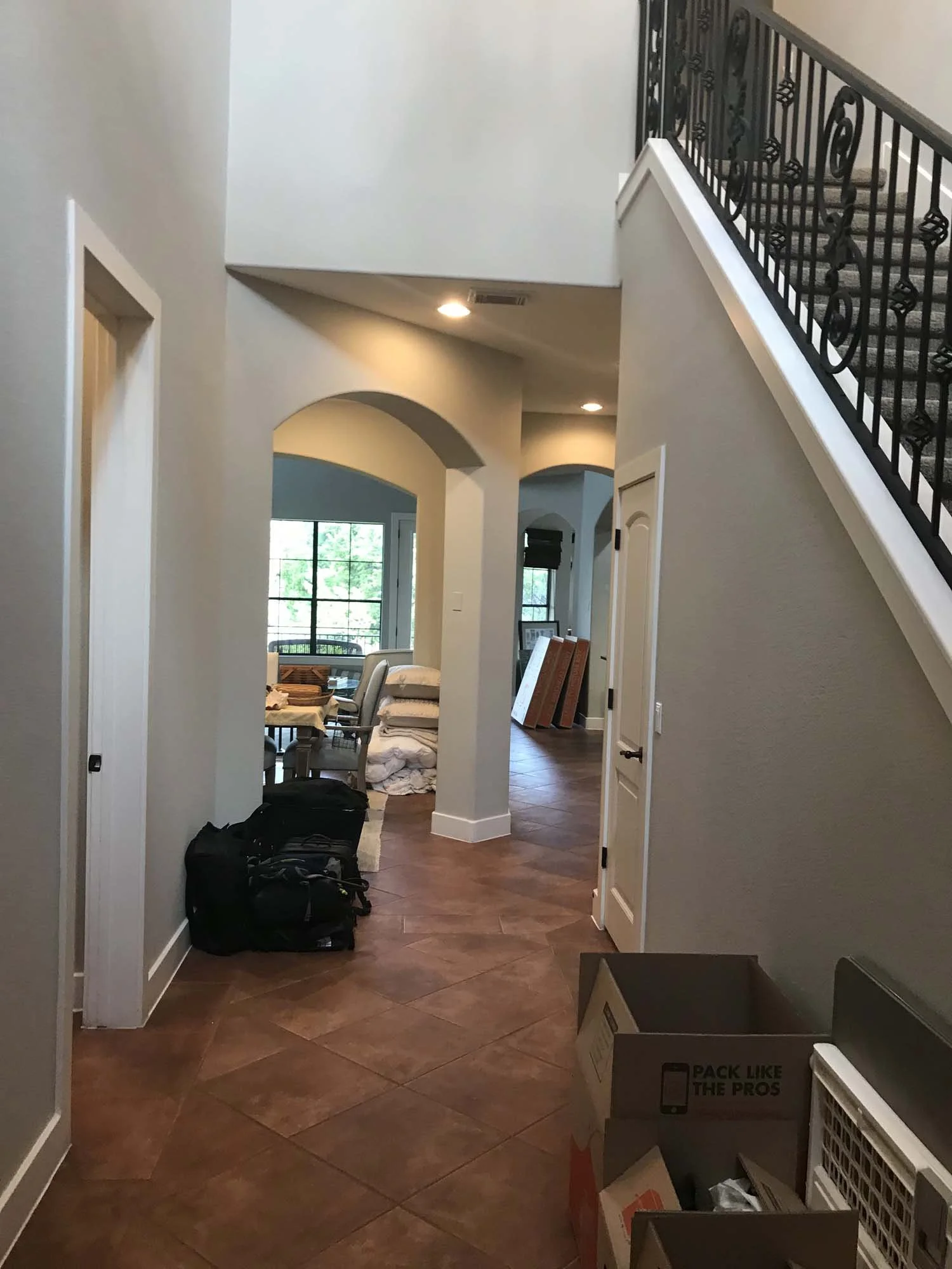 Interior view of a house with tile flooring, featuring stairs with a black wrought iron railing on the right, an open doorway, and a window letting in natural light. There are boxes and bags on the floor, and a stack of pillows or cushions.