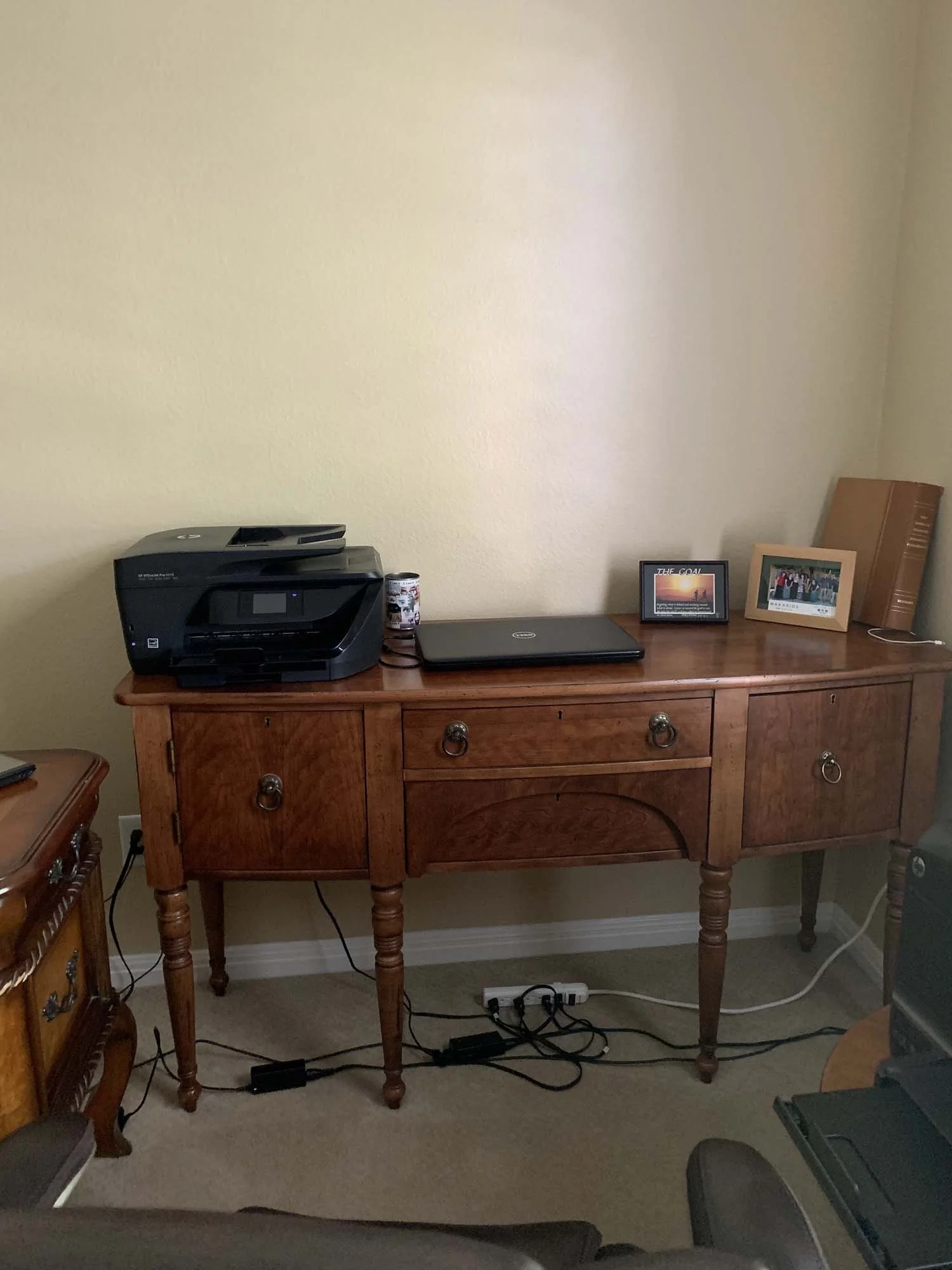 A wooden side table with various items on top, including a black printer, a closed laptop, two framed photographs, and a large book. Multiple power cords are visible underneath, plugged into a power strip.
