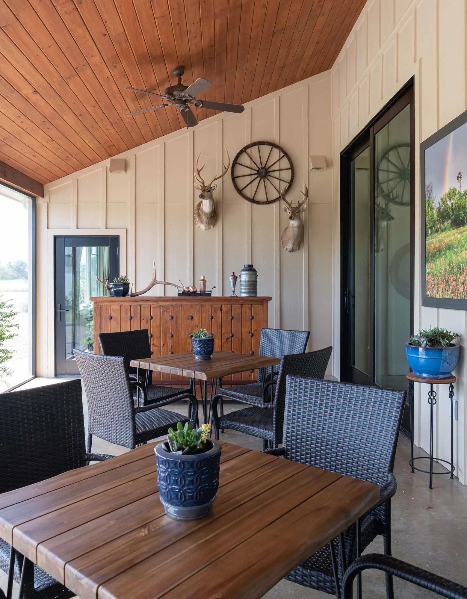 Indoor patio with wooden ceiling, beige walls, four black wicker chairs around two wooden tables, potted plants, deer head mounts, wagon wheel decor, ceiling fan, sliding glass doors, and a television.