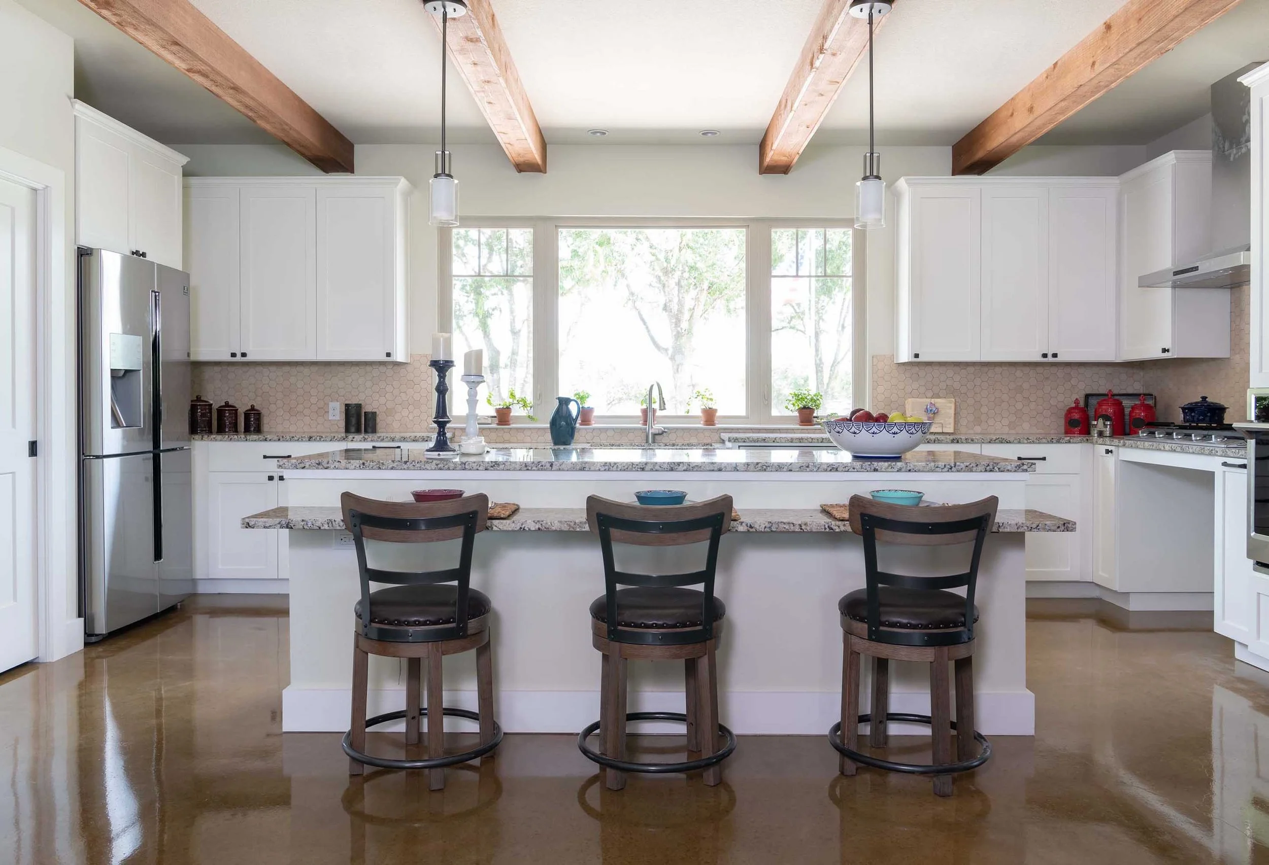 Modern kitchen with white cabinets, granite countertops, three black and wooden bar stools, large window with tree view, pendant lights, and various kitchen items.