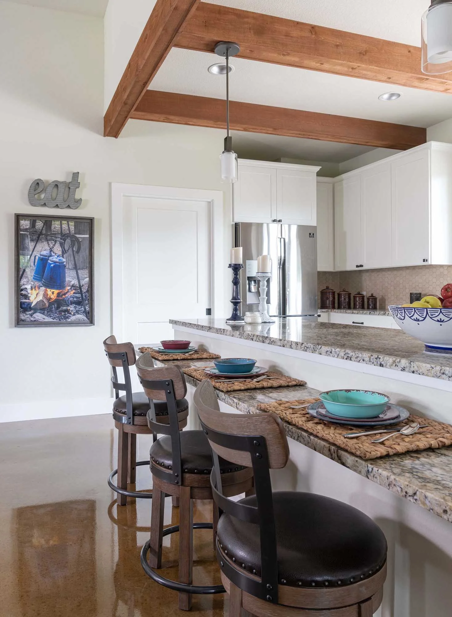 A kitchen with white cabinets and a granite countertop. Four barstools with dark wood and leather seats are positioned along the bar. The wall has a framed picture and a decorative sign that says 'eat.' The ceiling has wooden beams and a hanging ligh