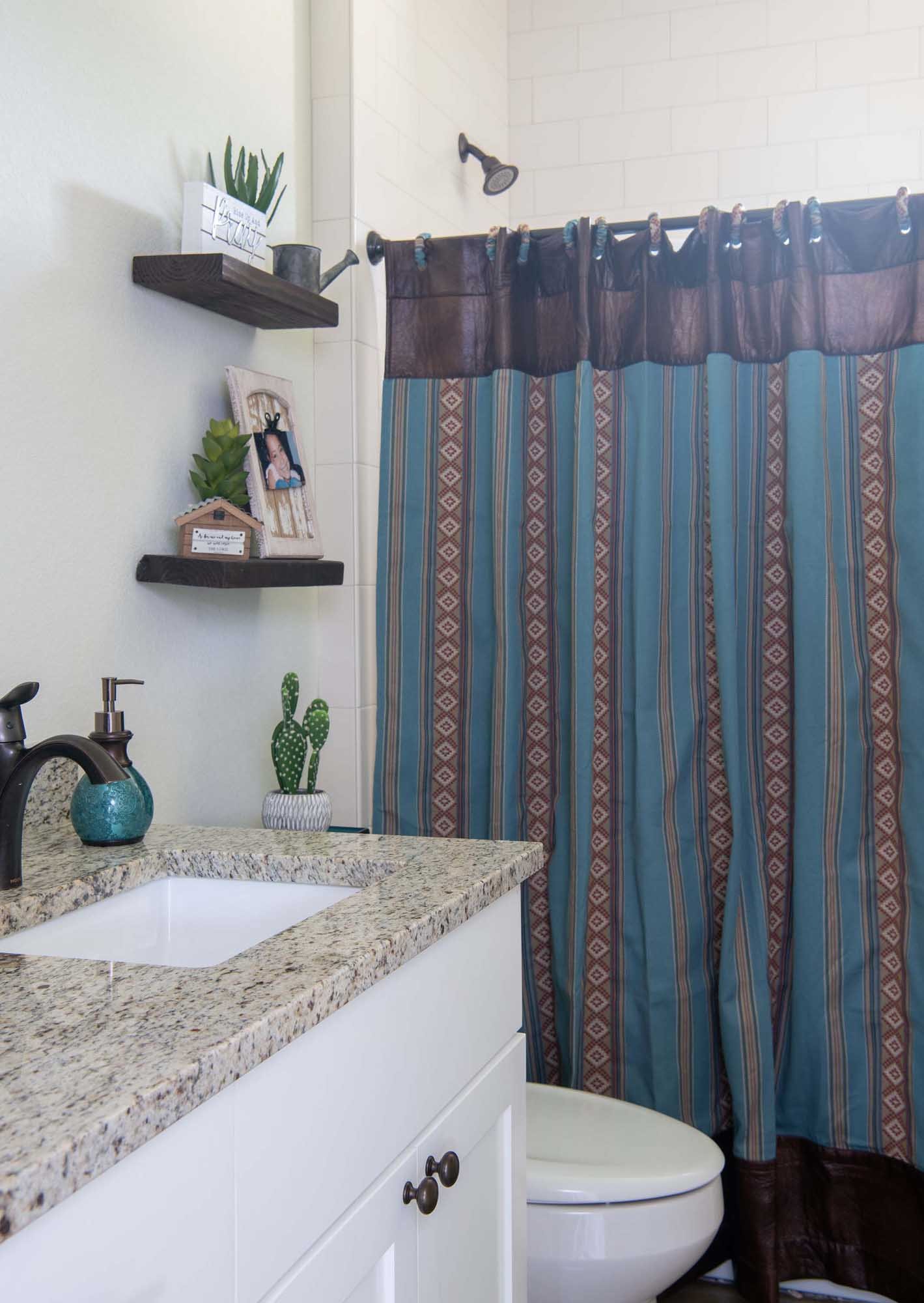 Bathroom with a granite countertop sink, a toilet, a shower with a striped curtain, and wall-mounted shelves decorated with plants, a framed photo, and small decor items.
