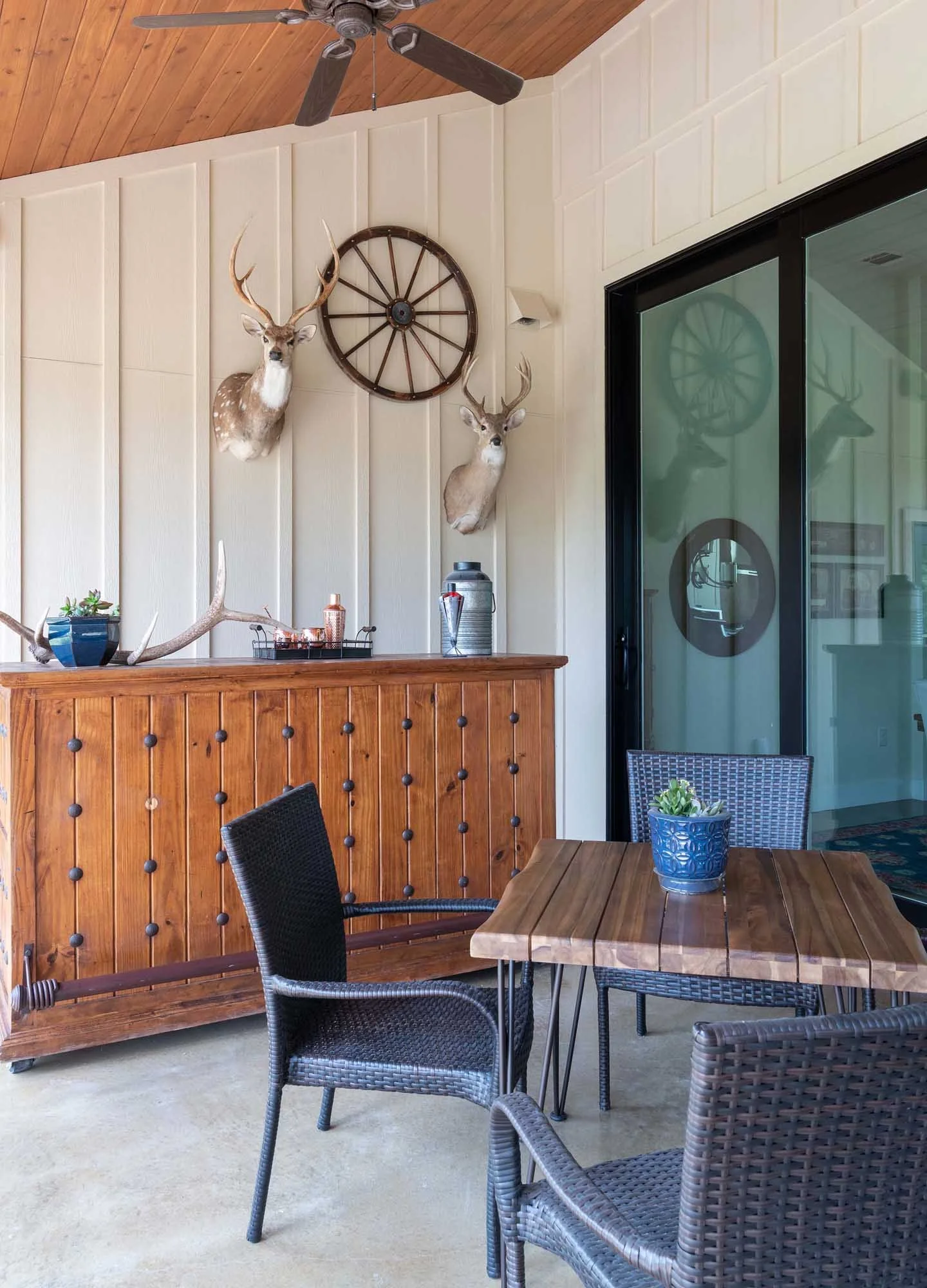 Indoor patio area with a wooden table and wicker chairs, a wooden sideboard, a glass sliding door, and two mounted deer heads with a wagon wheel on the beige paneled wall.