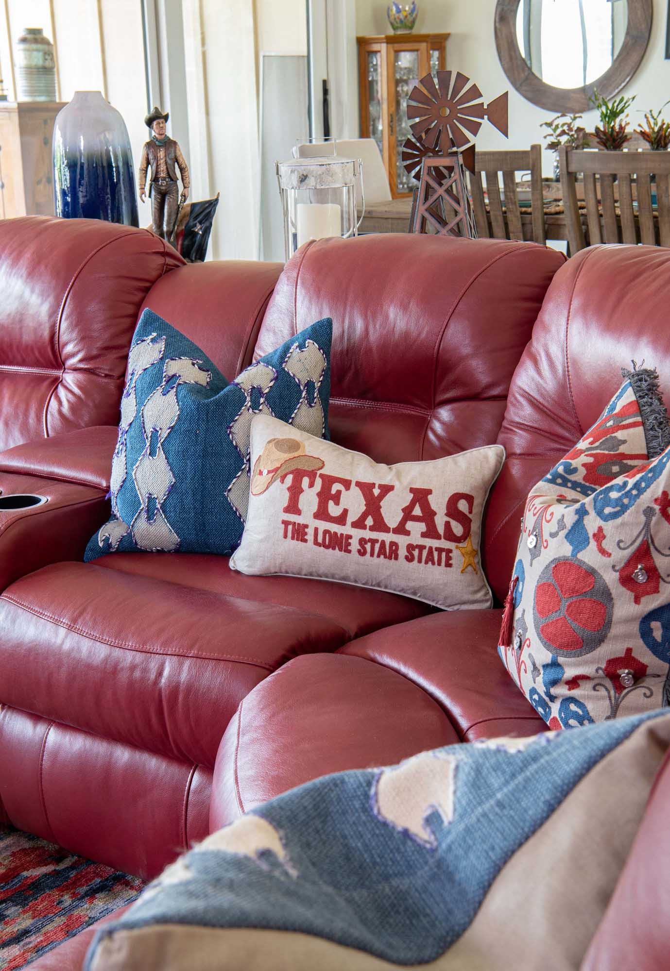 Living room with a red leather sofa decorated with Dallas Cowboys and Texas-themed pillows. The background shows a dining area with a windmill sculpture, a wooden cabinet, and decorative items.