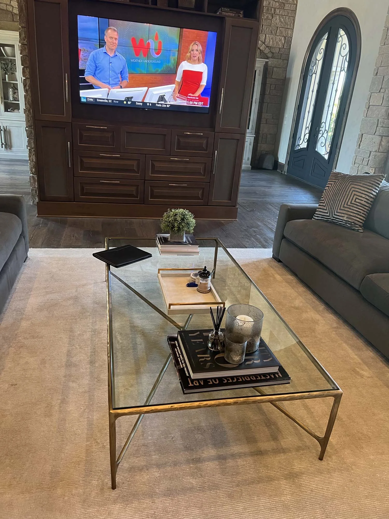 Living room with a glass-top coffee table holding books, candles, and decorative items, a TV wall with a weather forecast, and a door with decorative glass panels.