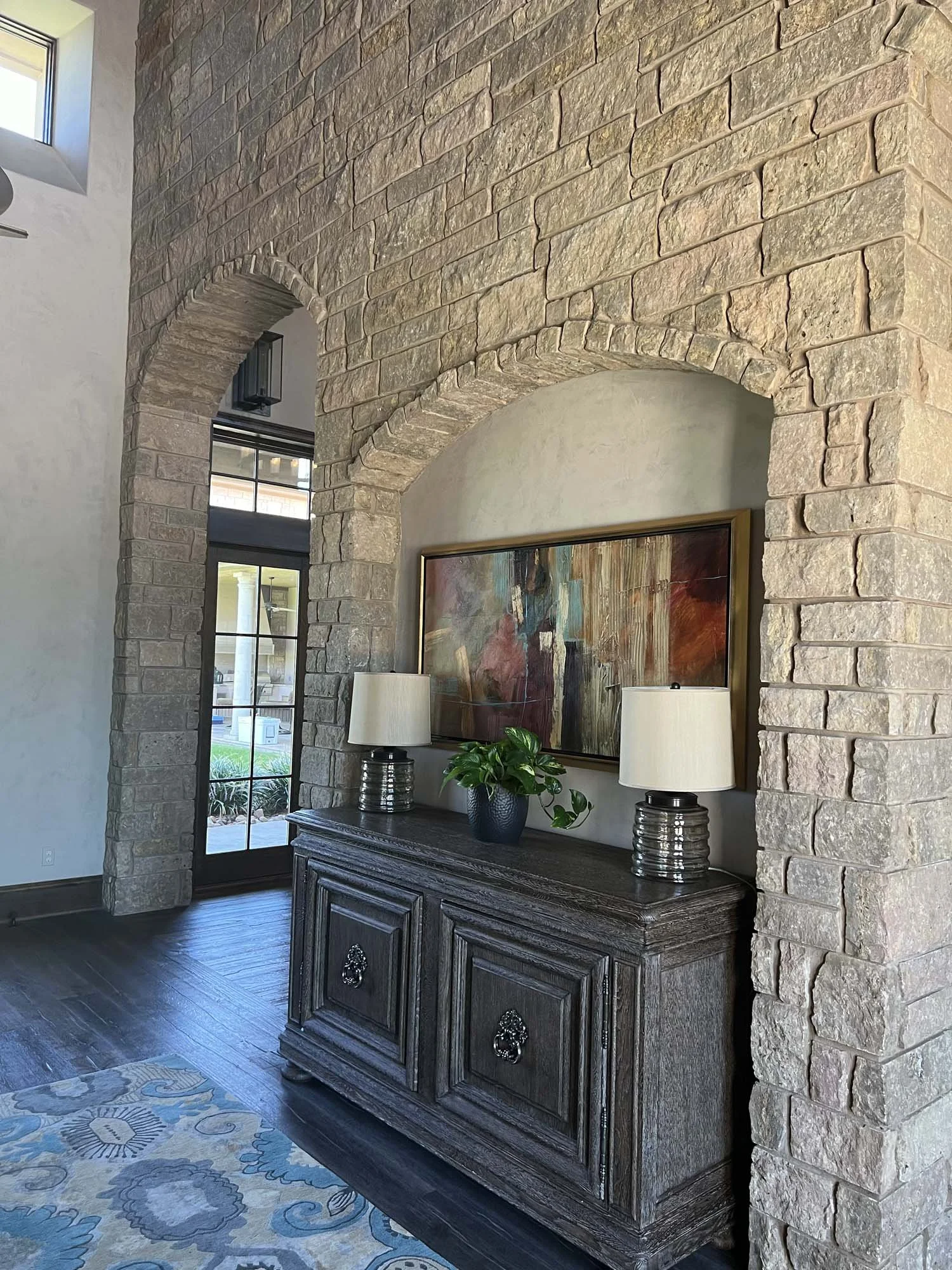 Interior of a room with a stone wall featuring two arches, a wooden sideboard with decorative handles, two table lamps with white shades, a potted plant, and an abstract painting mounted above the sideboard.