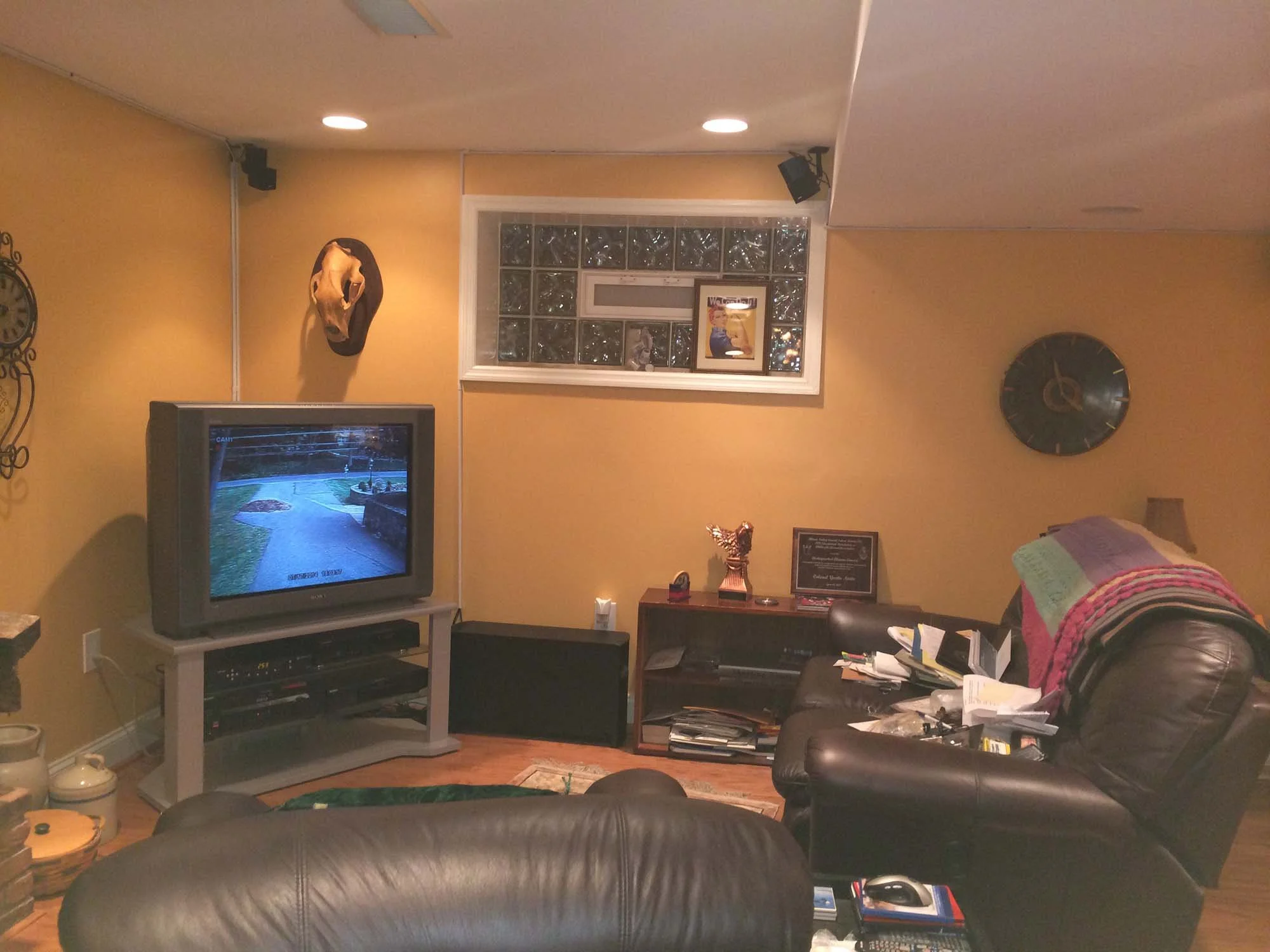 Living room with yellow walls, a television displaying a neighborhood scene, a framed picture on the window, and a leather armchair with a blanket and clutter on it.