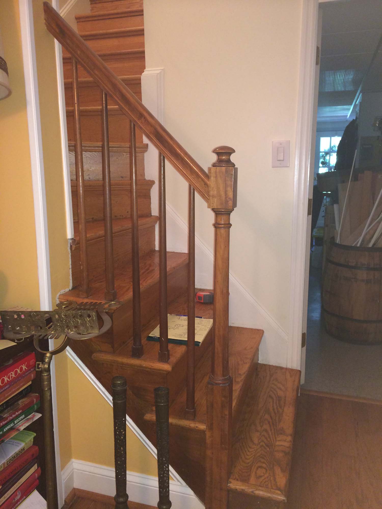 Wooden staircase with spindles and newel post, walls painted yellow and cream, doorway to another room with visible furniture and some tools.