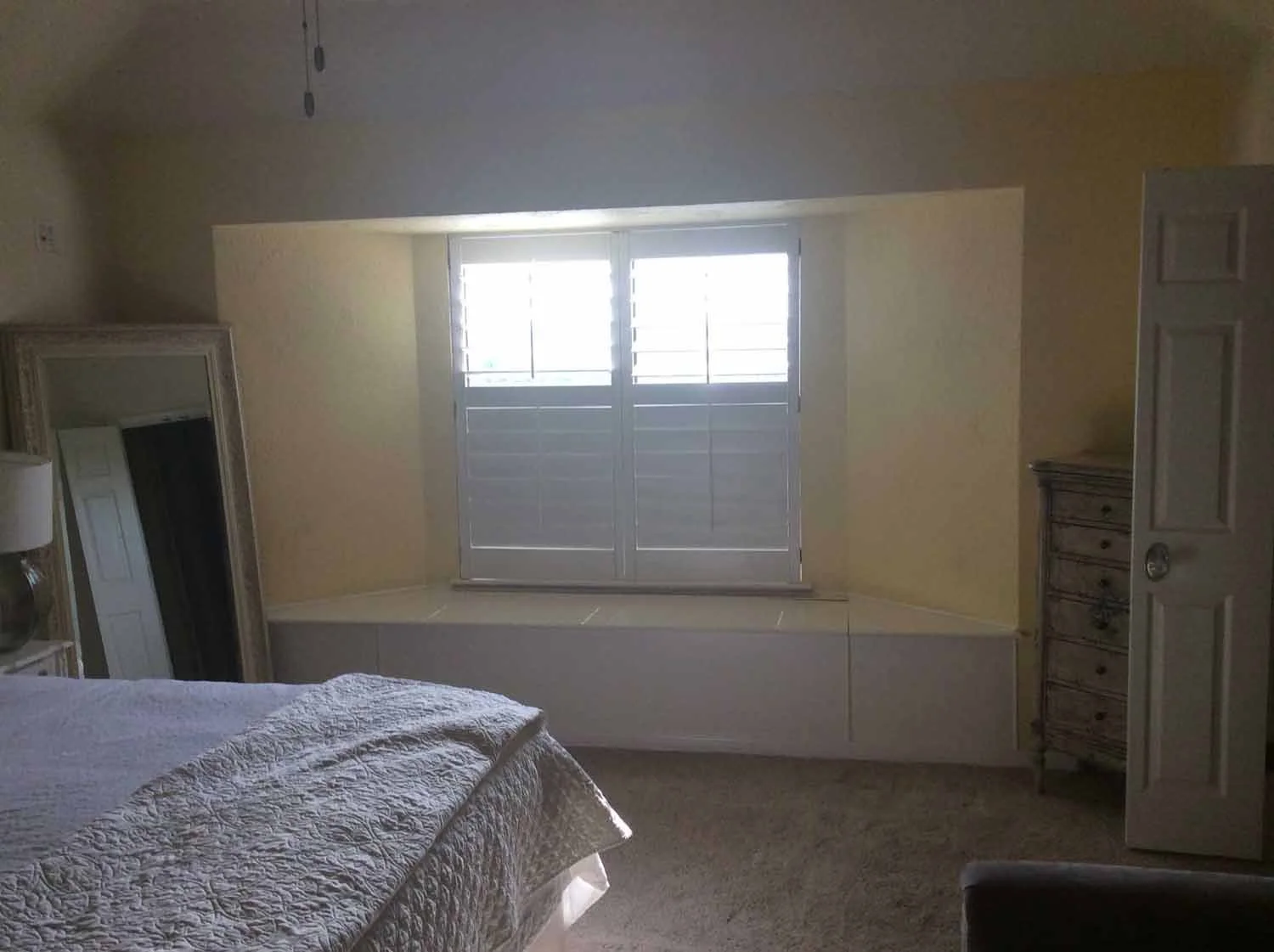 A bedroom with a bed covered in a white quilt, a piano with a mirror on top, a wooden dresser, and a window with white shutters.