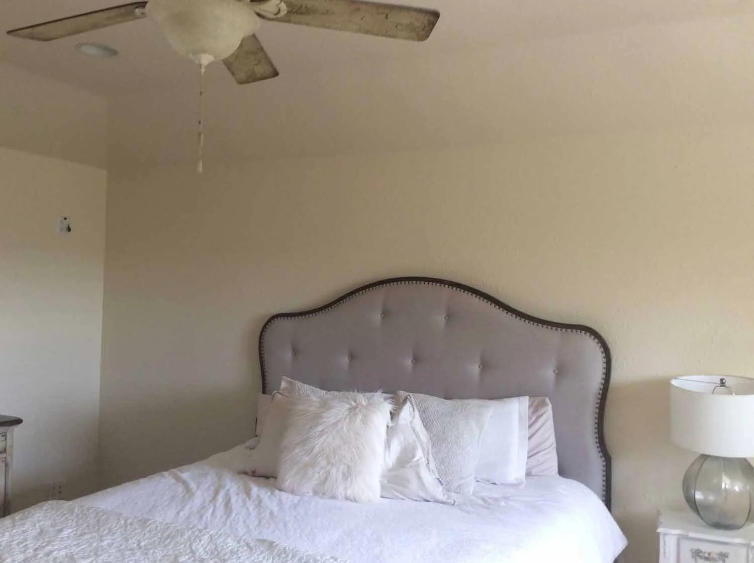 A bedroom with a bed having a light-colored upholstered headboard, white bedding, and pillows including a furry white pillow. A nightstand with a table lamp is on the right side of the bed. A ceiling fan with a light is above.