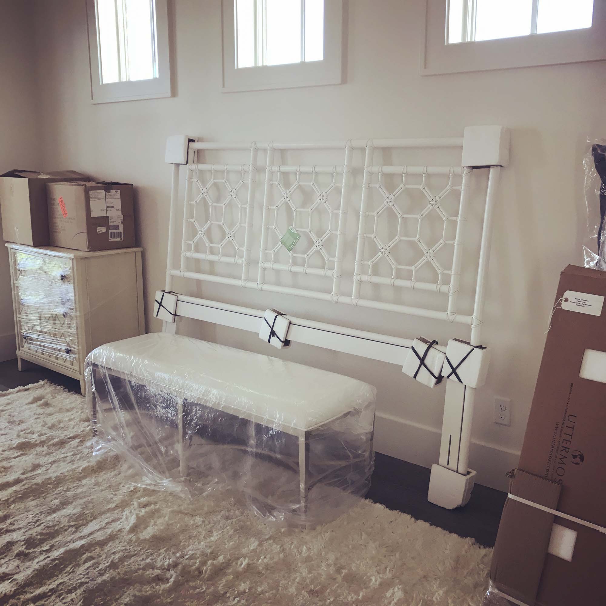 A white metal bed frame with a decorative headboard leaning against a wall, with a bench wrapped in plastic in front of it. There are boxed items on a dresser to the left and a large cardboard box on the right. The room has beige walls, three windows