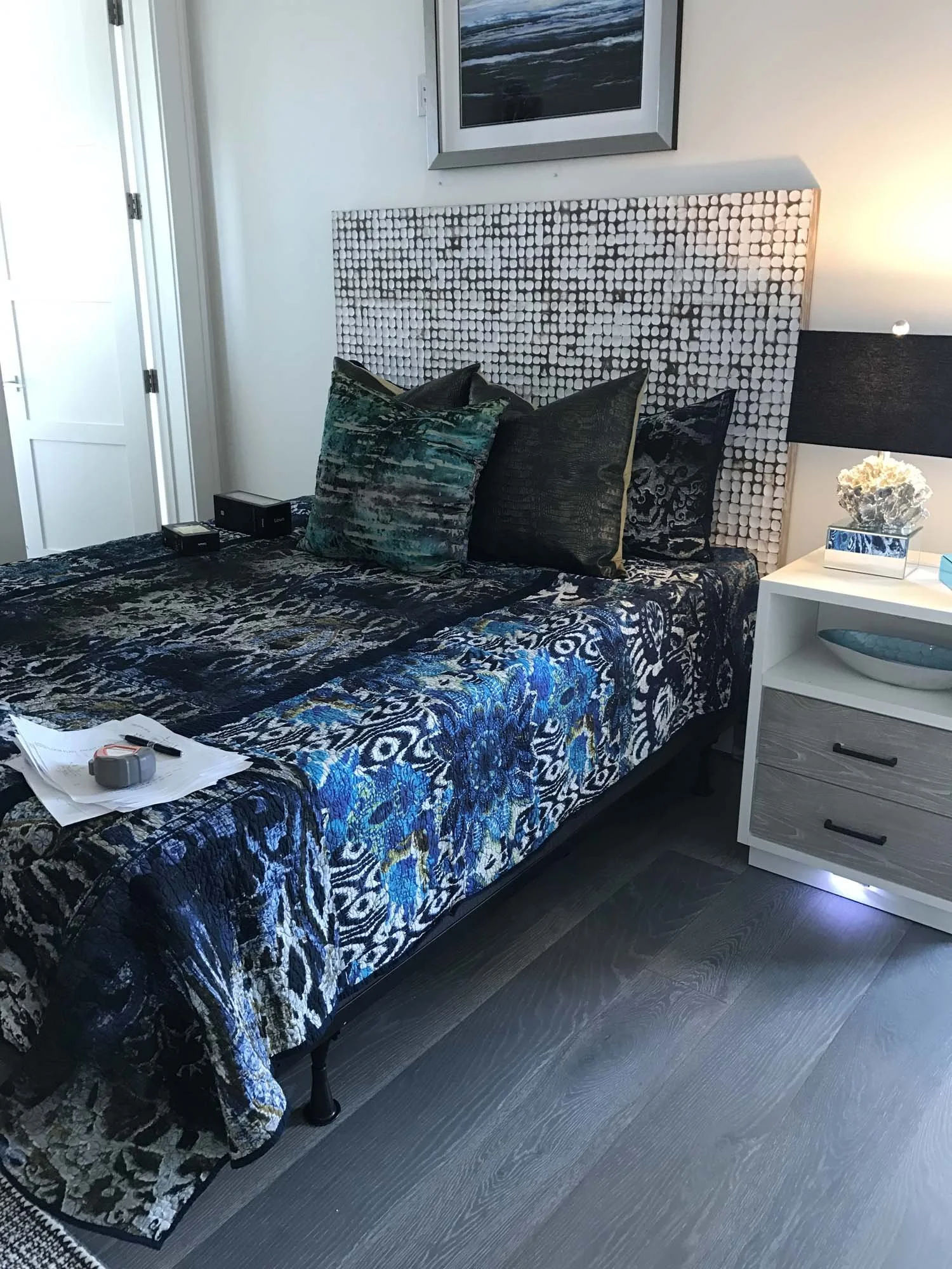 A hotel room with a bed featuring a black, blue, and white patterned bedspread and matching pillows. There is a white nightstand with a lamp, decorative items, and a drawer to the right of the bed. A framed artwork hangs above the headboard.